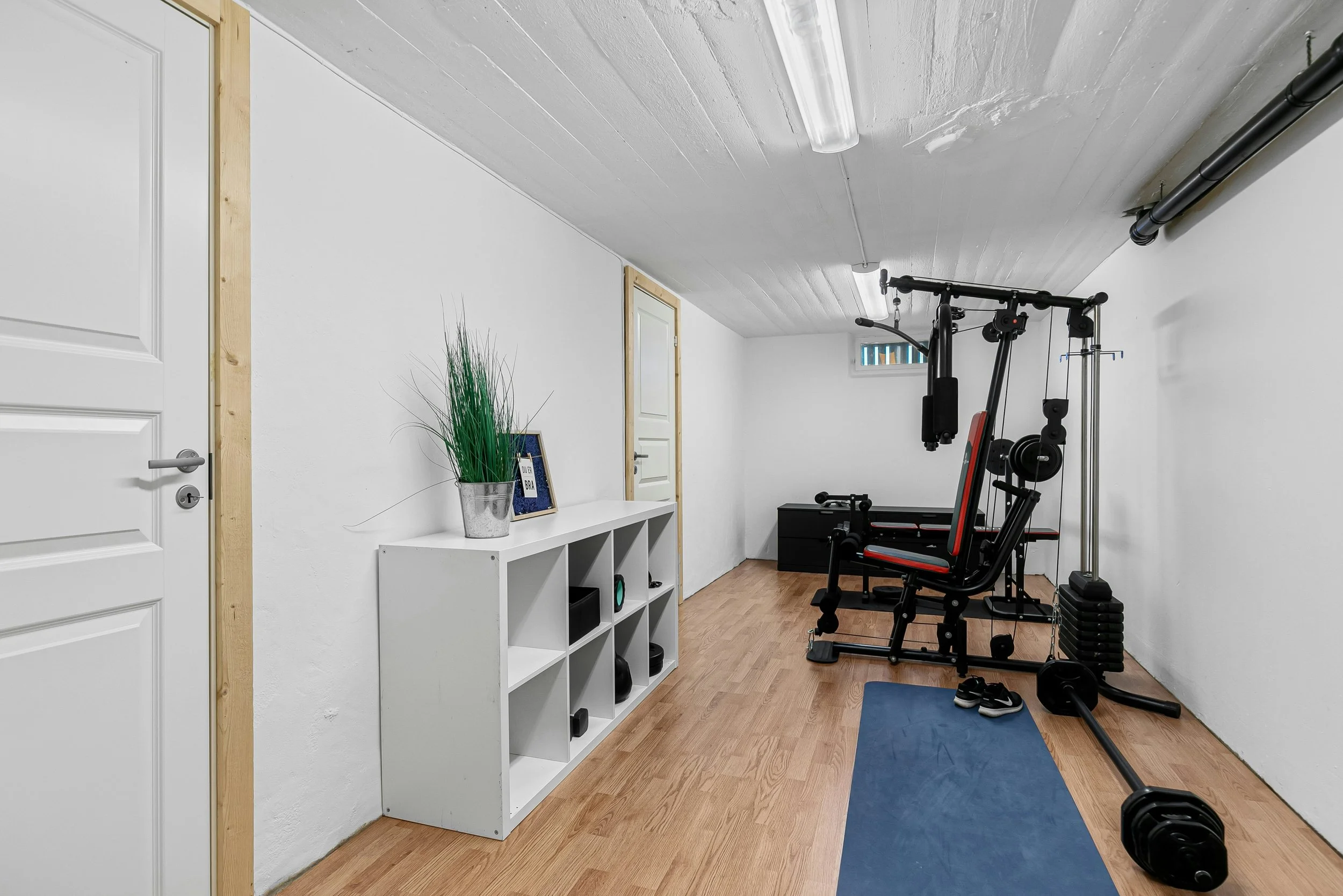 Home gym with exercise equipment including a multi-function weight machine, a barbell on the floor, and weight plates, inside a room with white walls, wooden floor, a white shelf with decor, and a small window.