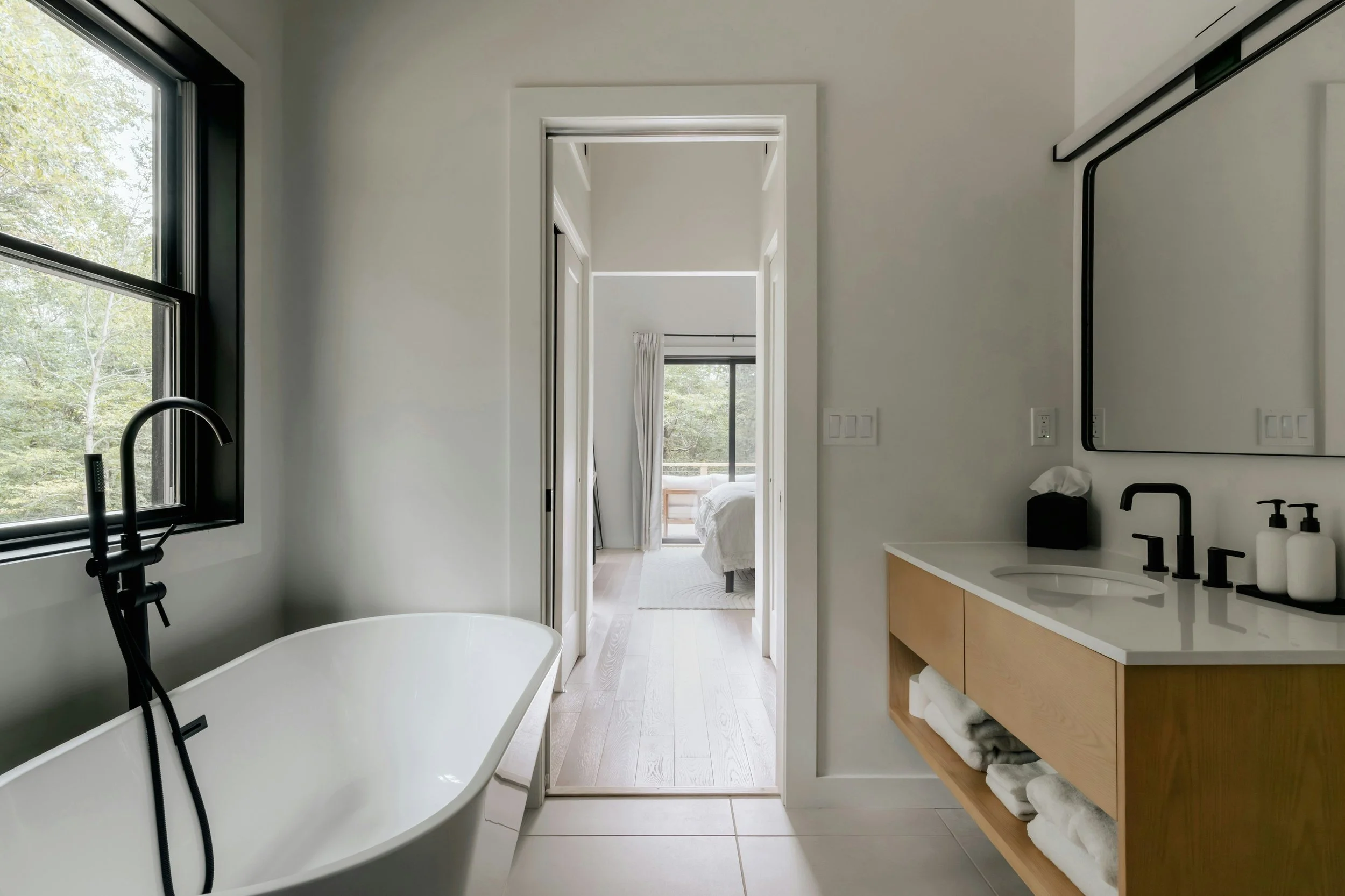 Modern bathroom with white walls, black-framed window, freestanding bathtub, wooden vanity with open shelf, white towels, black faucet, and mirror, view into bedroom with large window and bed.