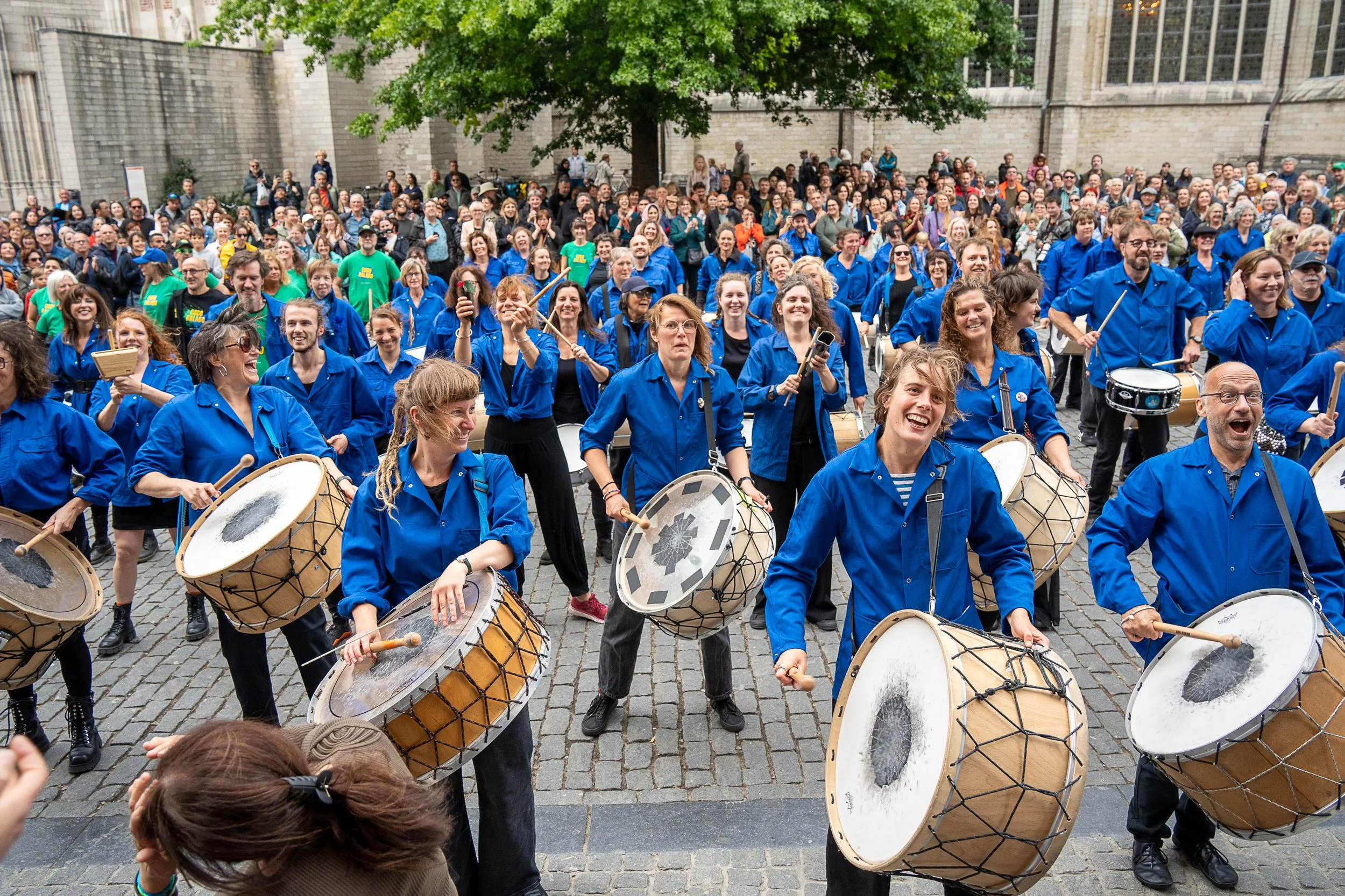Een grote groep percussionisten in blauwe hemden speelt enthousiast op straat voor een glimlachend publiek.