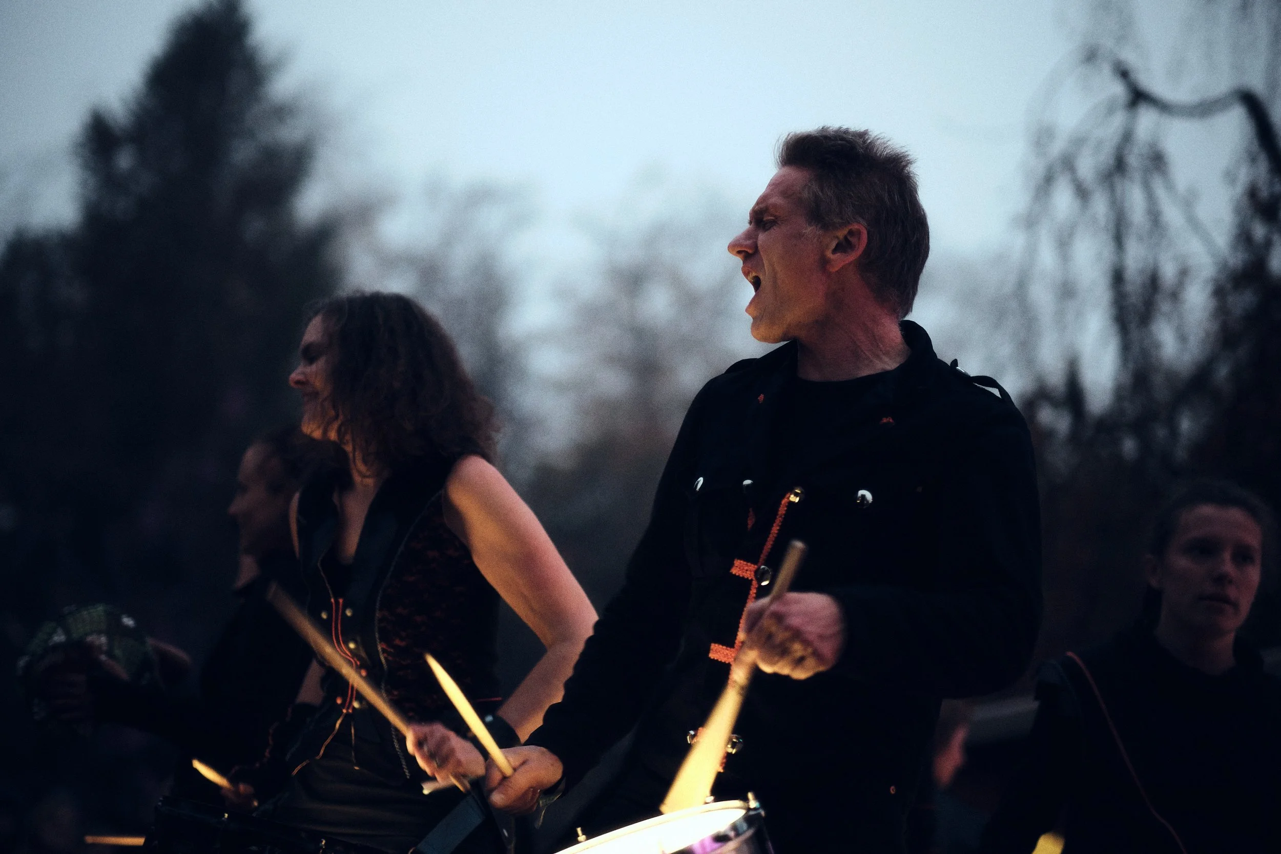 Jo Zanders, oprichter van De Batterij, speelt vol energie op zijn trom tijdens een buitenoptreden in de schemering, omringd door medepercussionisten.