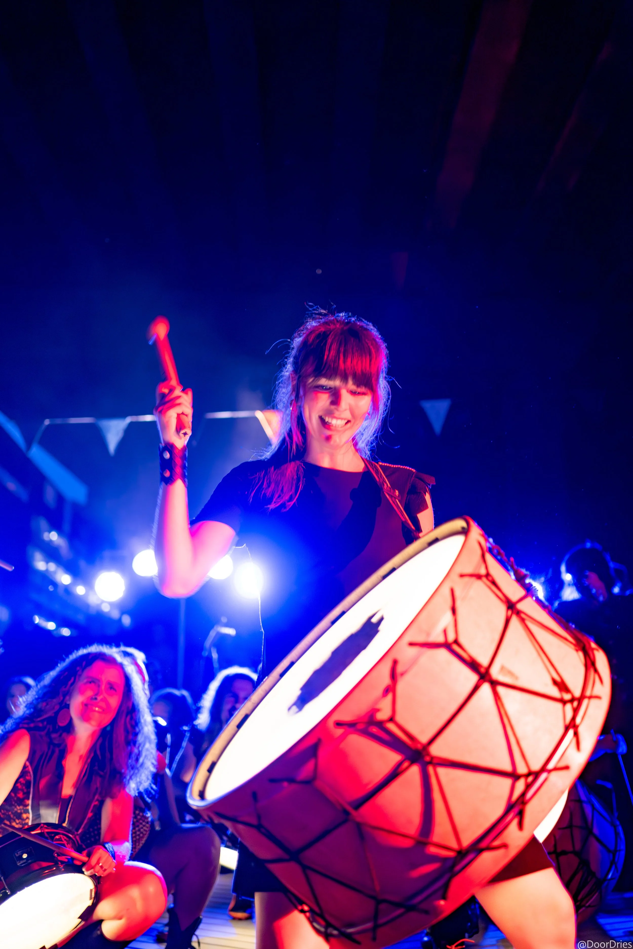 Vrouw die trommelt tijdens een live optreden, omringd door muzikanten in blauw licht.