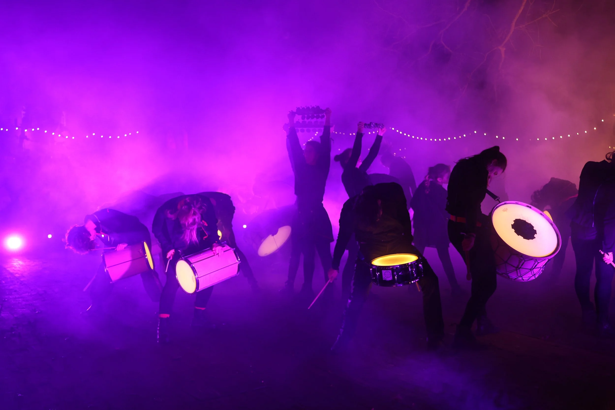Een groep drummers treedt op in de buitenlucht, omgeven door paarse en roze nevel en sfeerverlichting.