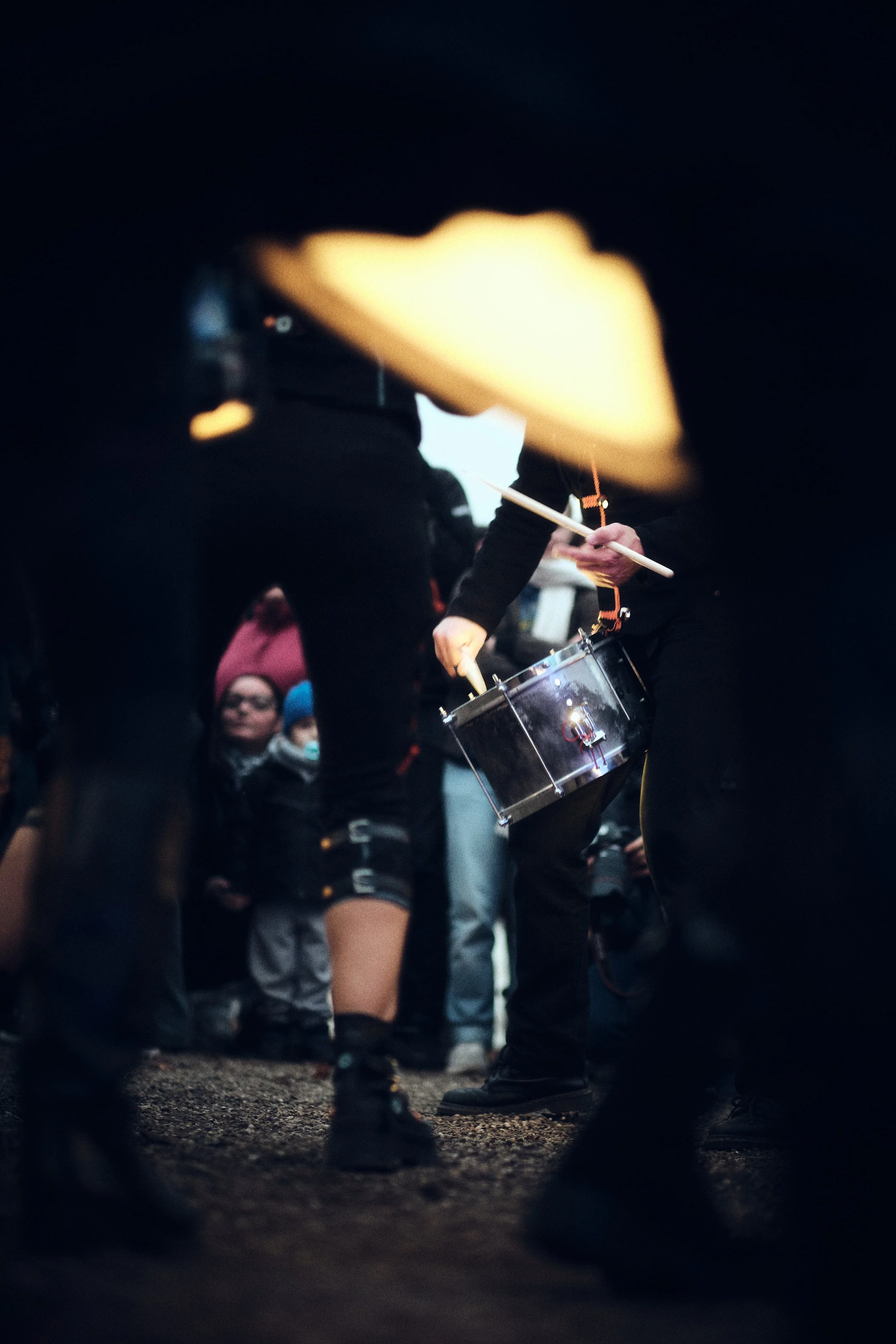 Close-up van een drummer die in de buitenlucht speelt, omringd door toeschouwers en medeperformers in donkere kleding.