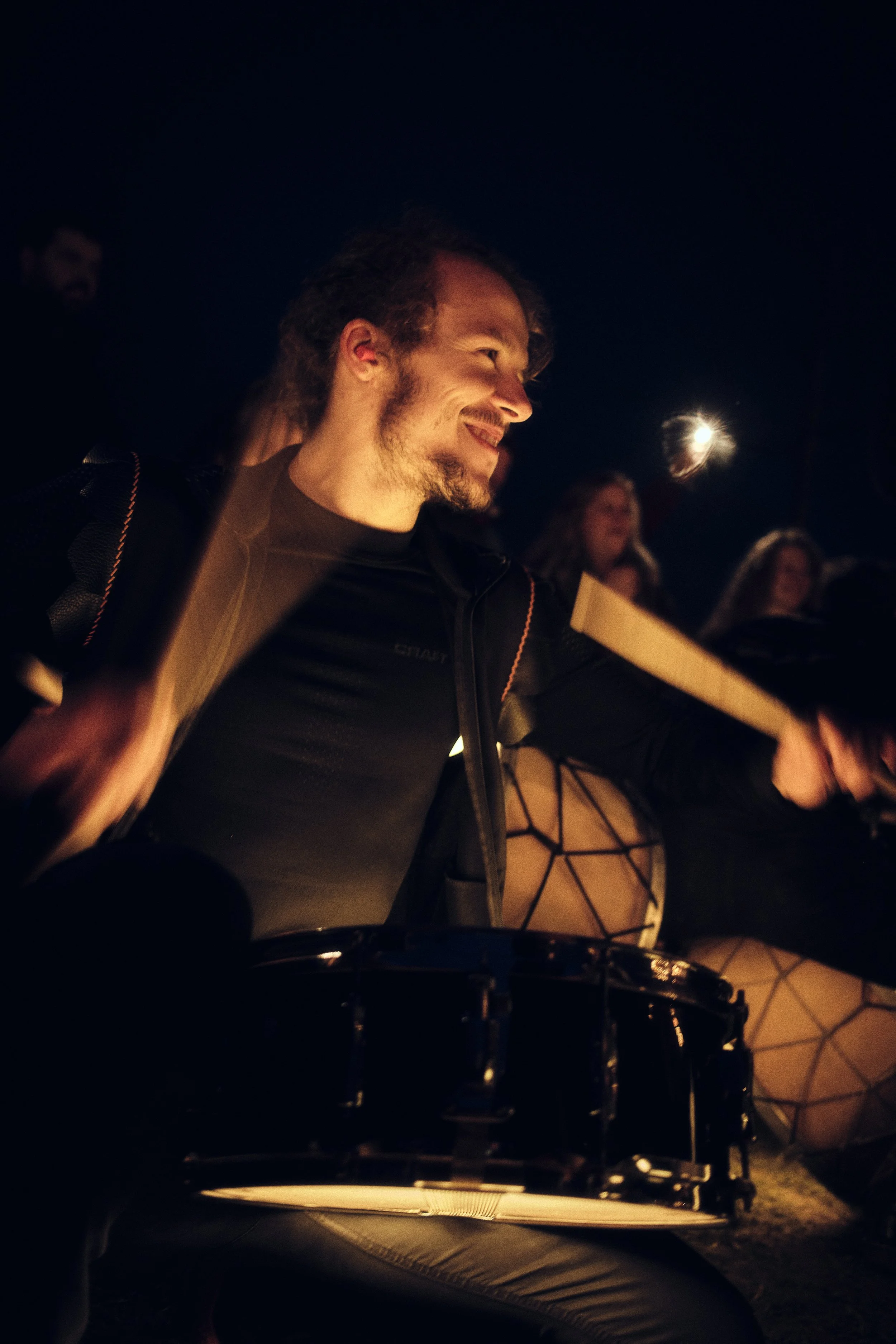 Drummer speelt enthousiast op een trommel tijdens een nachtelijk optreden, warm verlicht door omgevingslicht.