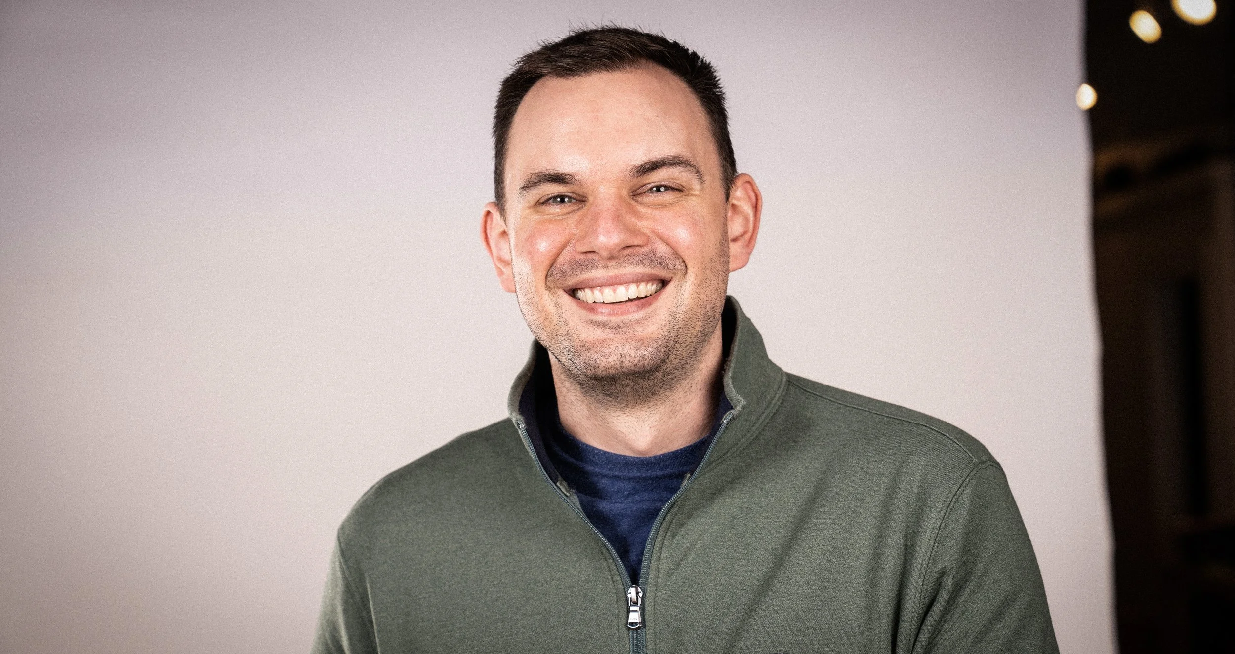 A smiling man with short dark hair, wearing a green zip-up jacket over a dark blue shirt, standing in front of a plain, light-colored background.