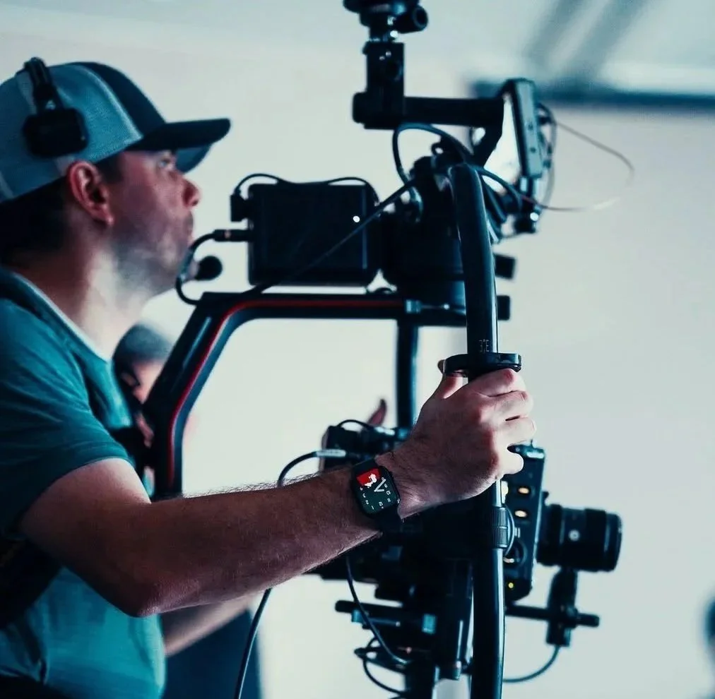 A videographer wearing a baseball cap operating a professional camera setup with a monitor and other equipment.