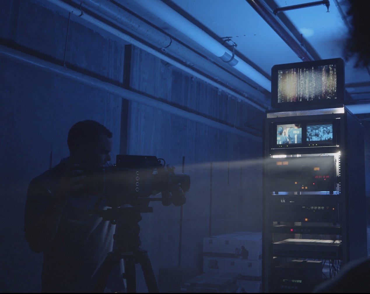 A person operating a professional video camera in a dark, blue-lit room with electronic equipment and monitors.