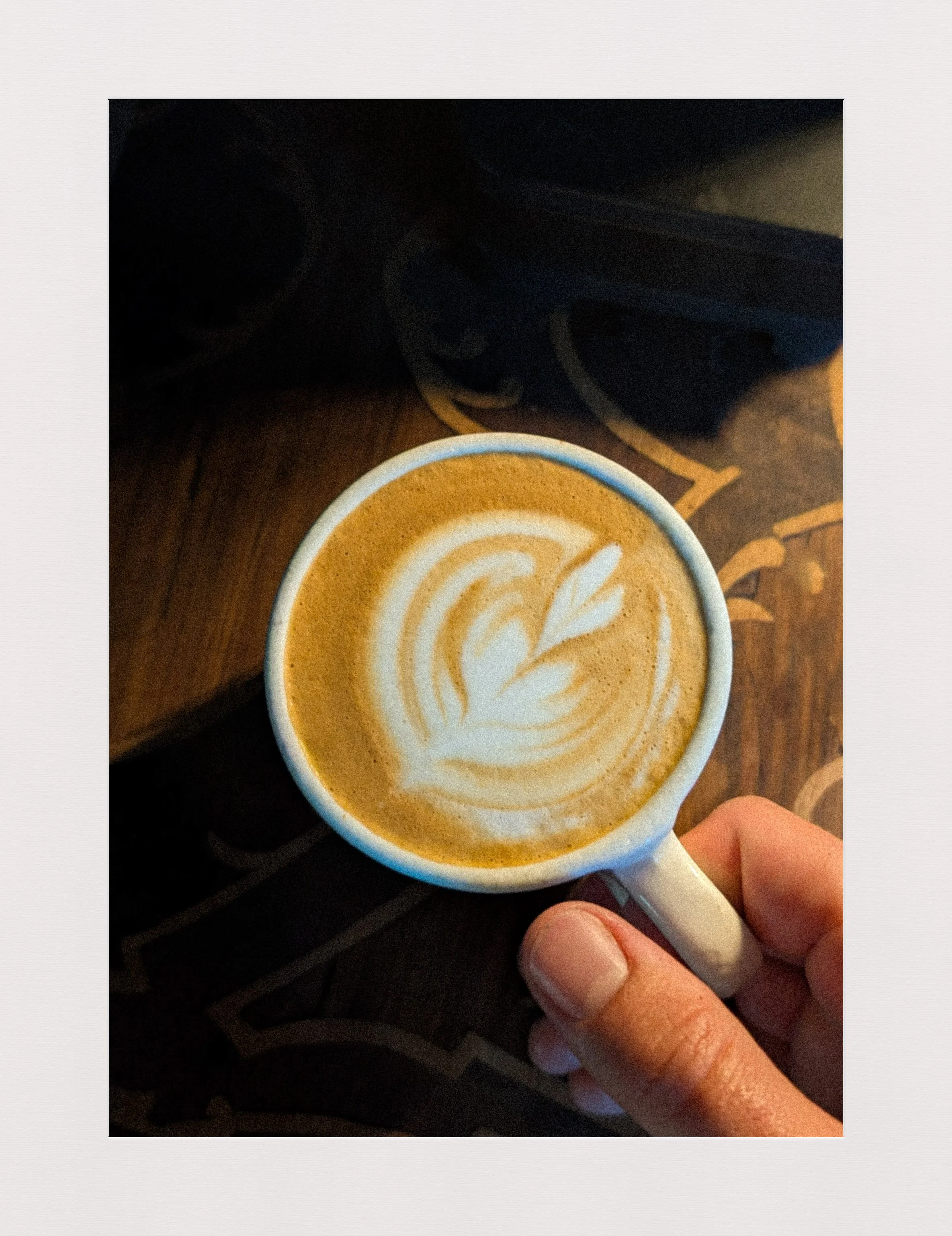 A hand holding a white coffee mug with latte art on top, displaying a leaf design, on a dark wooden table.