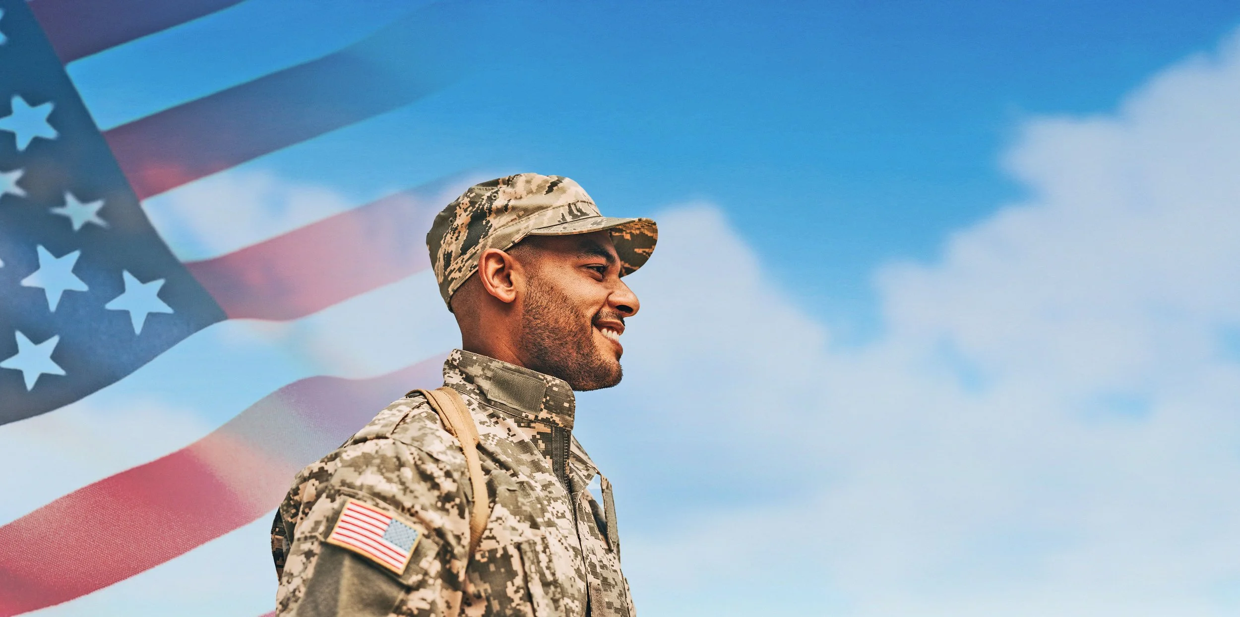 A smiling soldier in camouflage uniform and cap standing outdoors with the American flag waving in the background against a blue sky with some clouds.