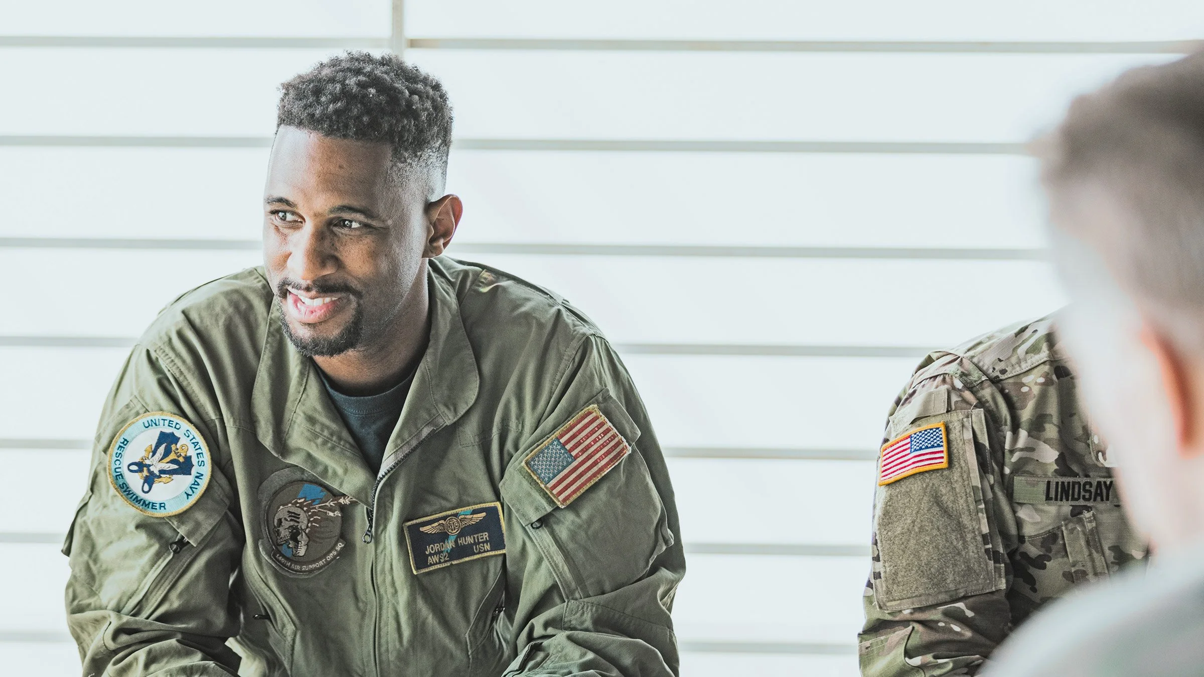 Two military personnel, one with a patch reading 'United States Navy Rescue Swimmer' and wearing an American flag patch, sitting and talking, while another in camouflage uniform with a patch reading 'Lindsay' is partially visible on the right, in a room with white horizontal paneling.