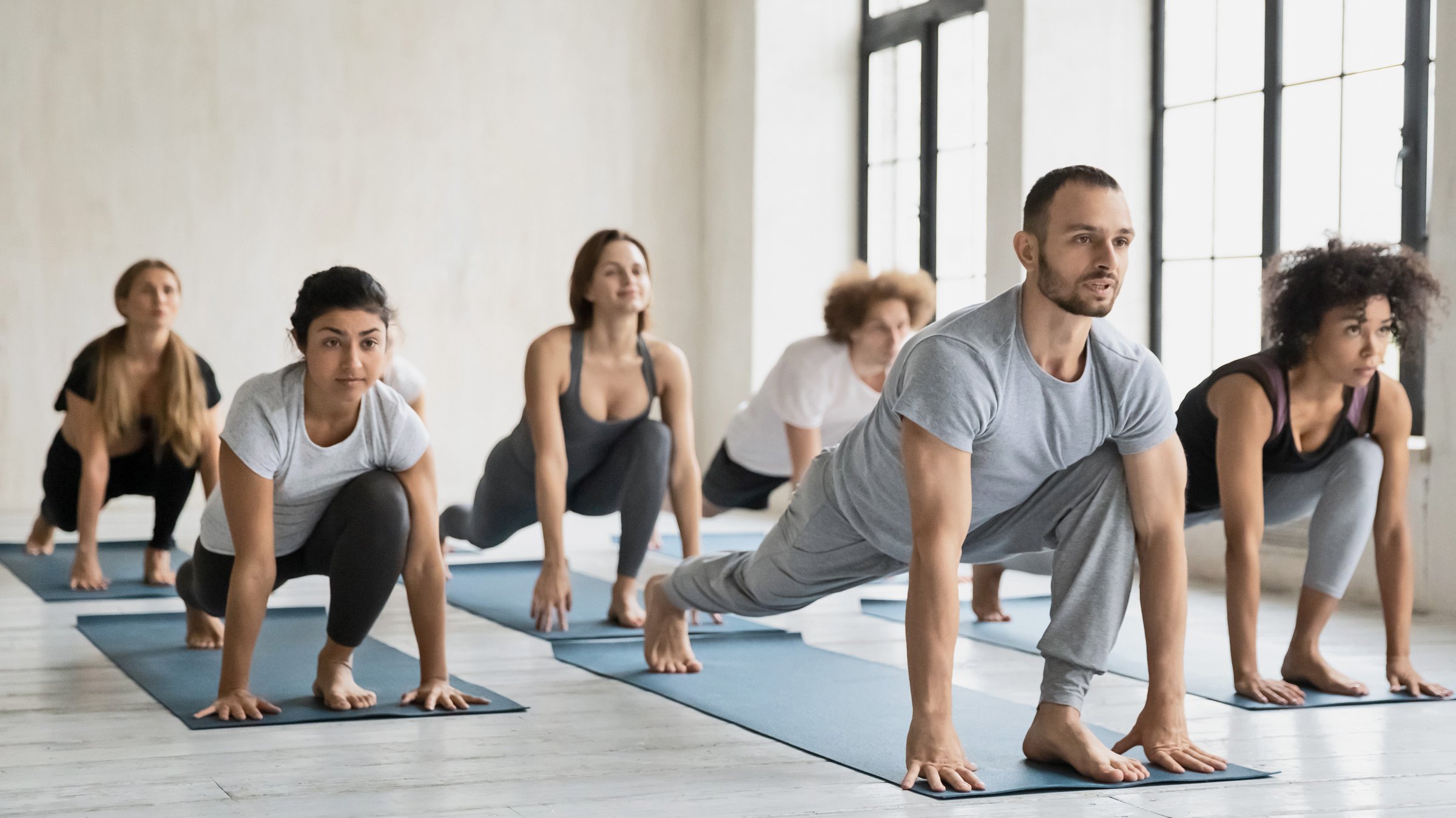 Group of diverse people practicing yoga in a spacious studio, performing a lunge pose on yoga mats near large windows.