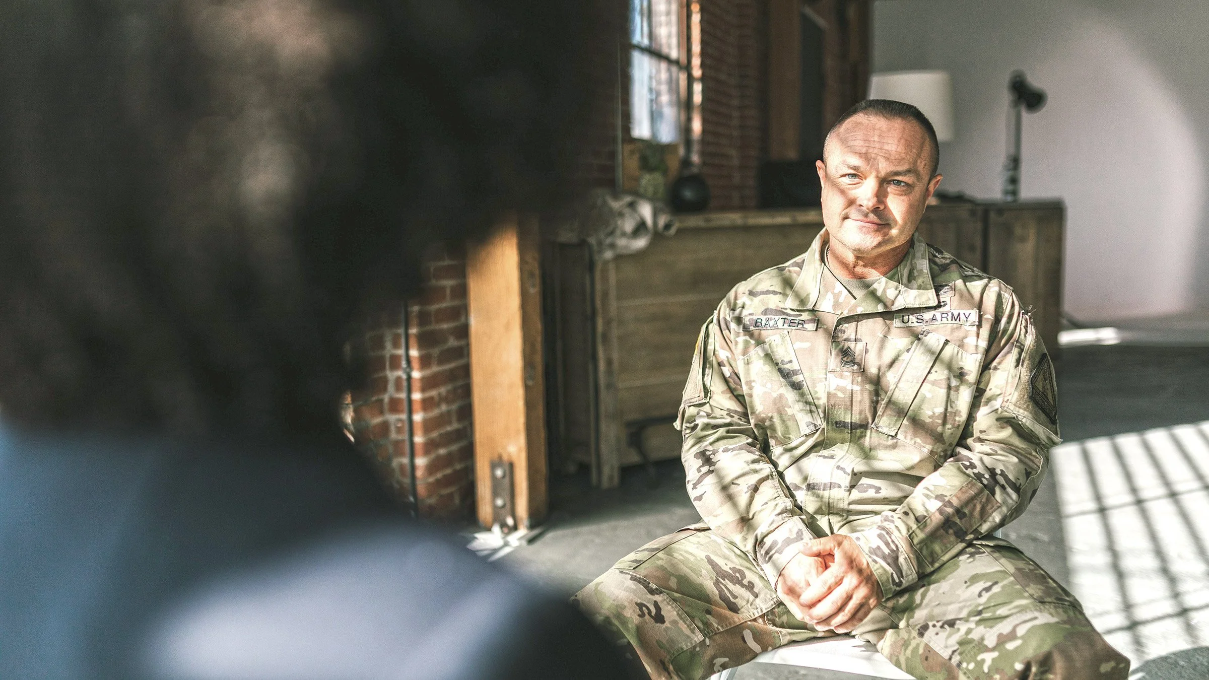 U.S. Army soldier in camouflage uniform sitting on a bench in a rustic room with brick walls, having a conversation, with sunlight streaming through a window.