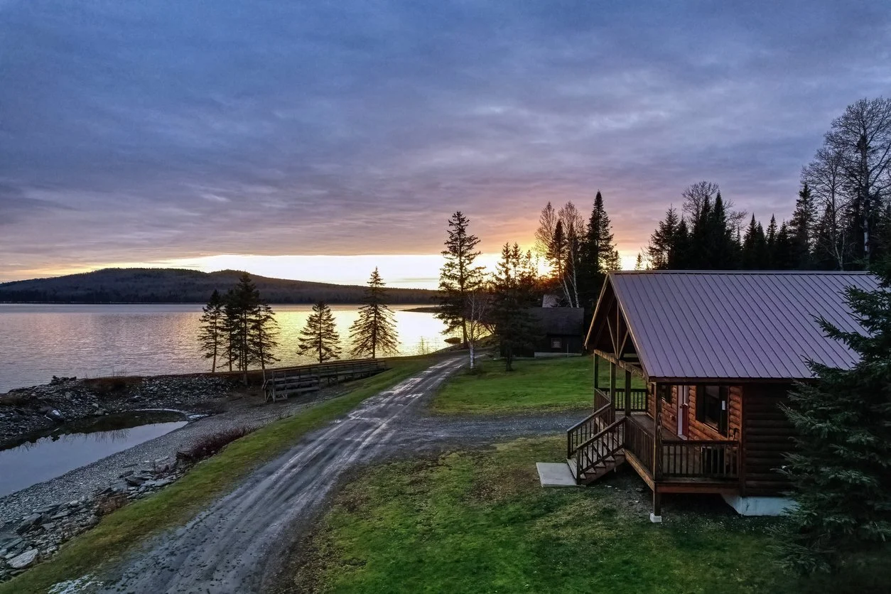 A scenic lakeside view at sunset with a cabin, trees, a dirt road, and a cloudy sky.
