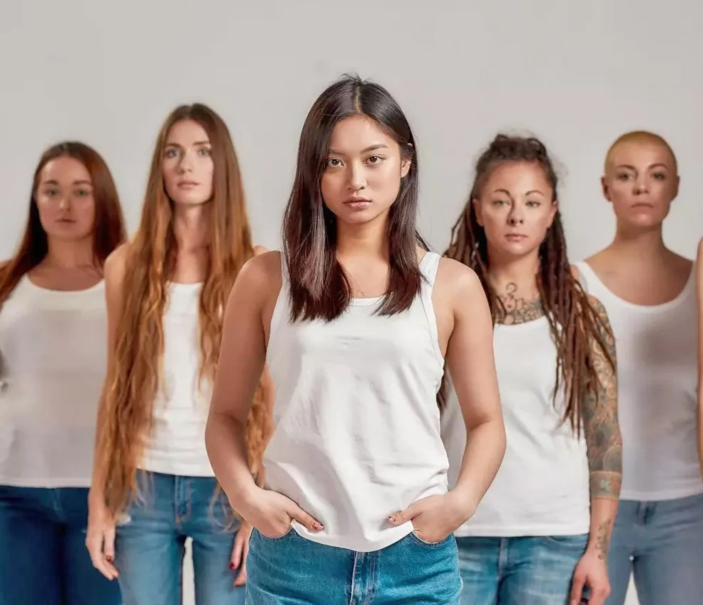 A diverse group of five women standing confidently with their hands in their pockets, wearing white tank tops and jeans, against a plain background.