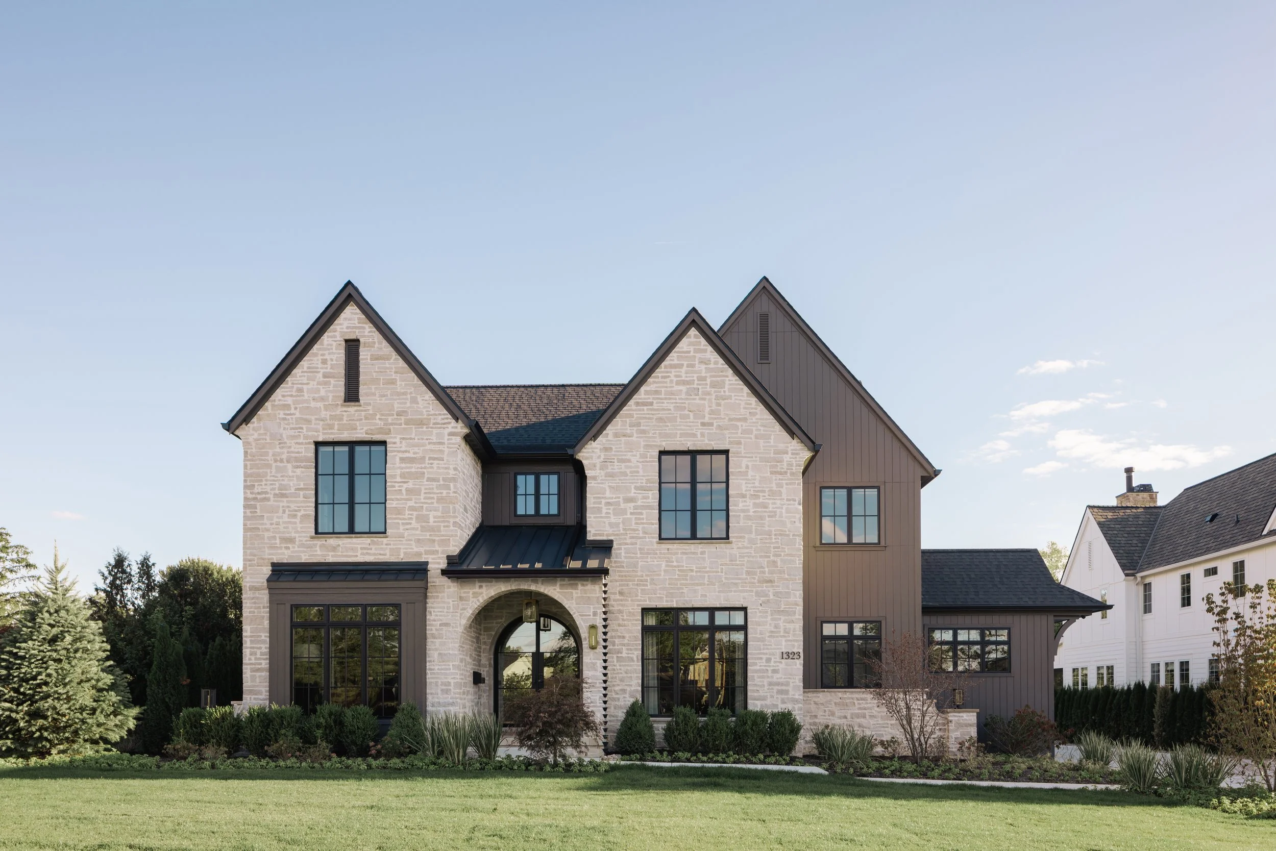 Front view of a modern house with neutral stone and vertical dark siding, with two gabled roofs and a central arched entryway with glass windows, surrounded by a well-kept lawn and small bushes.