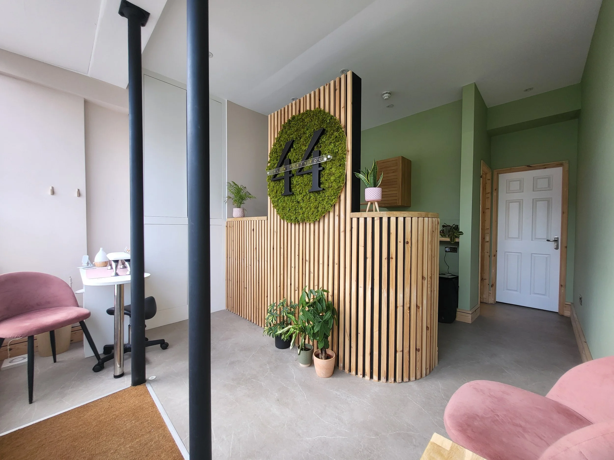Interior of a modern office or lounge area with a wooden slat reception desk, potted plants, a pink chair, and a large circular wall clock with greenery background.