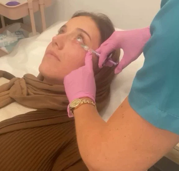 A woman lying in a hospital bed receiving a nasal injection from a healthcare worker wearing pink gloves and a blue uniform.