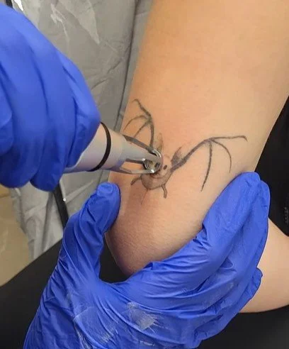 A tattoo artist is using a tattoo machine to ink a spider design onto a person's arm, with the person's arm supported by a gloved hand.