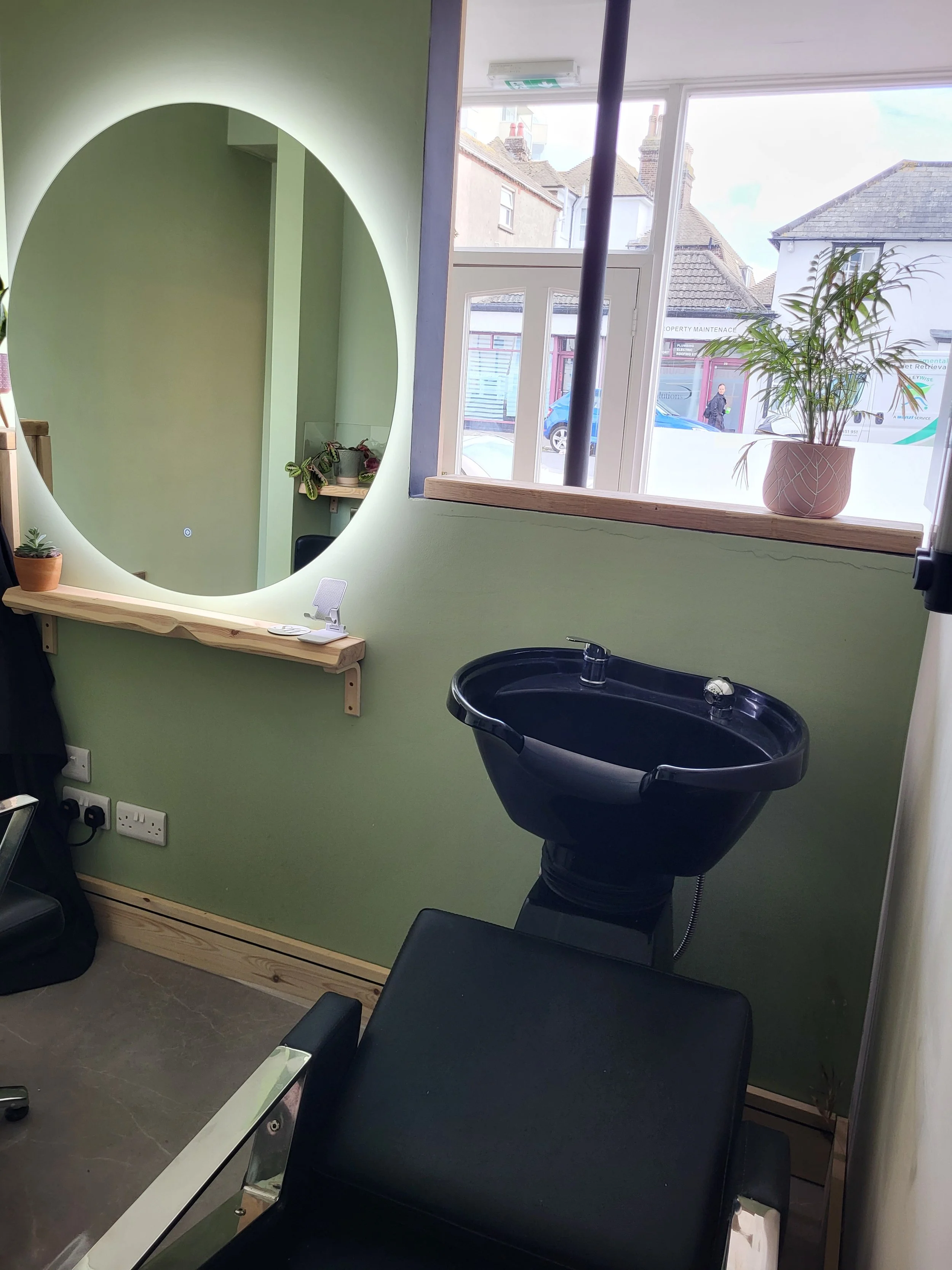 Inside a hair salon, with a black sink for hair washing, a large round mirror on a light green wall, a wooden shelf with potted plants, a window with a view of the street, and hair salon chairs.