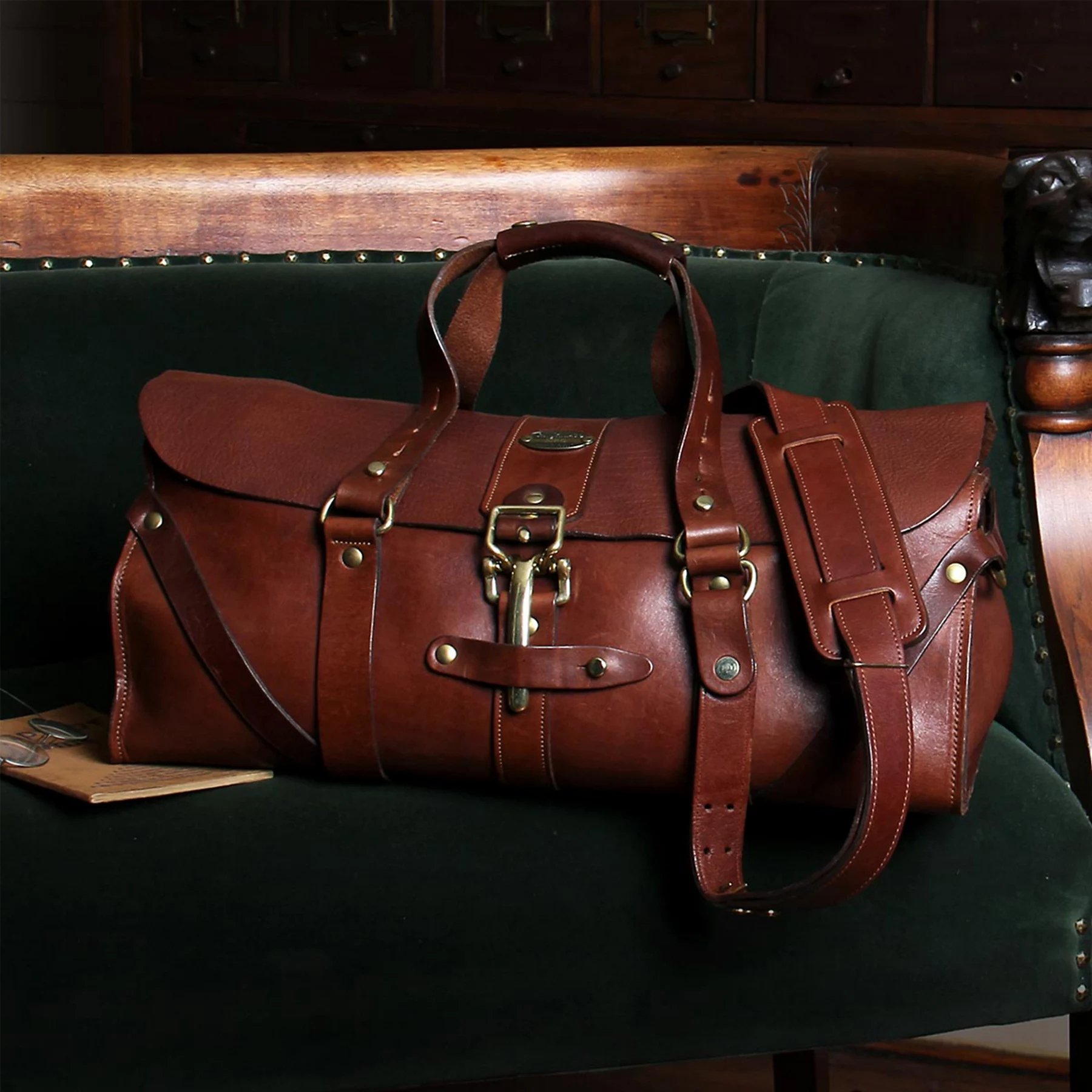 A brown leather duffel bag with brass hardware, resting on a dark green upholstered bench.