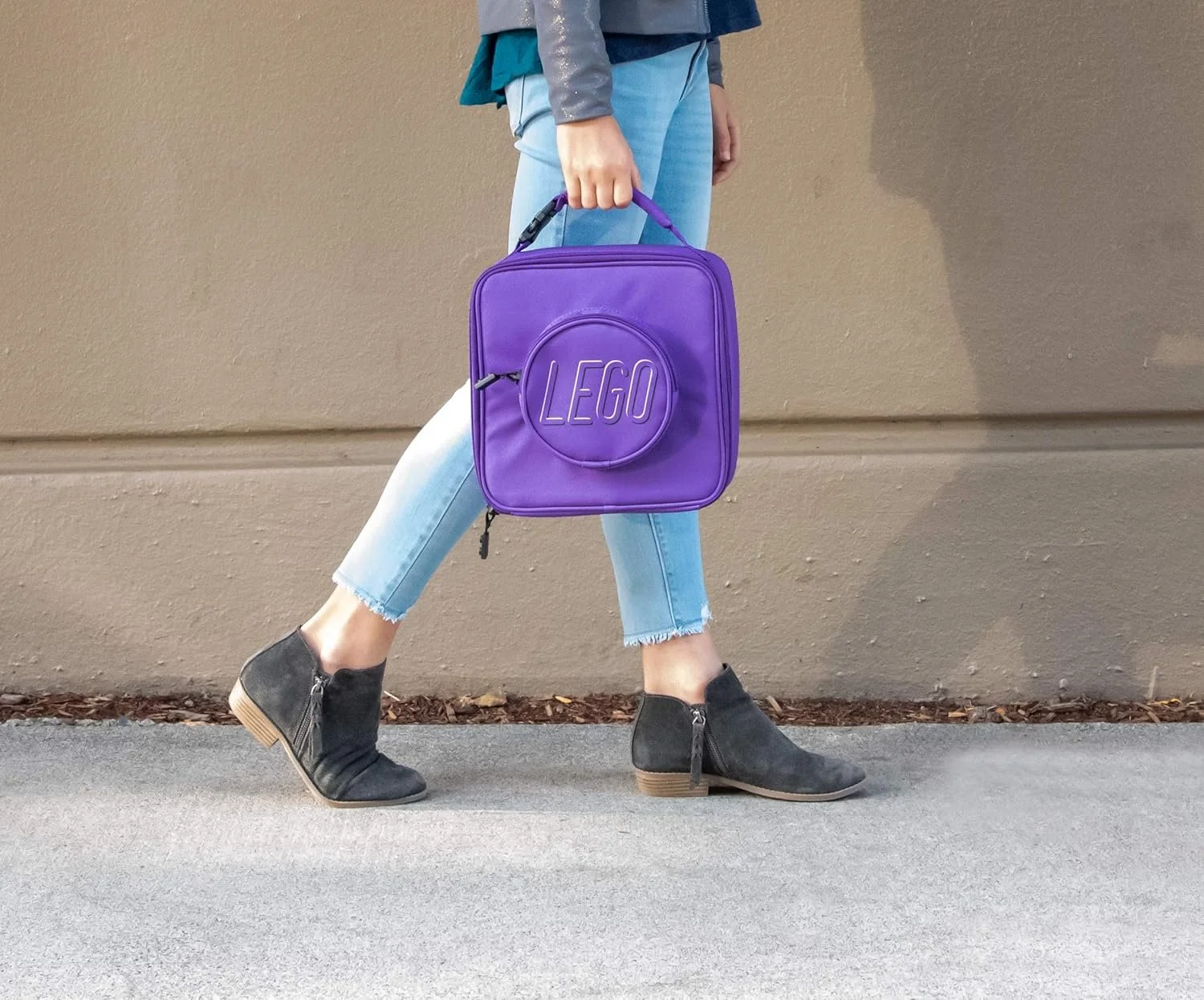 Person walking holding a purple LEGO-branded bag, wearing black ankle boots, light blue jeans, and a multicolored top, against a beige wall.