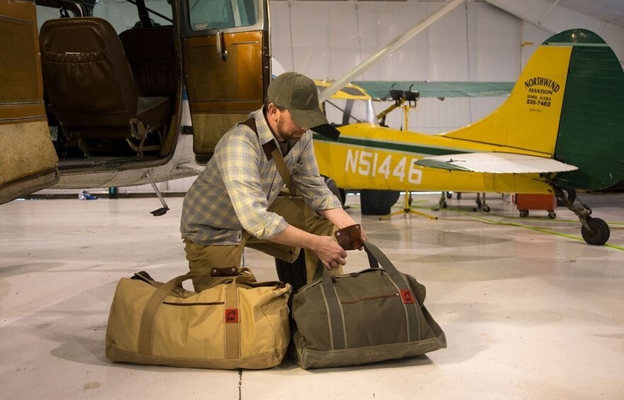 Man packing travel bags in airplane hangar with vintage yellow and green airplane in background.