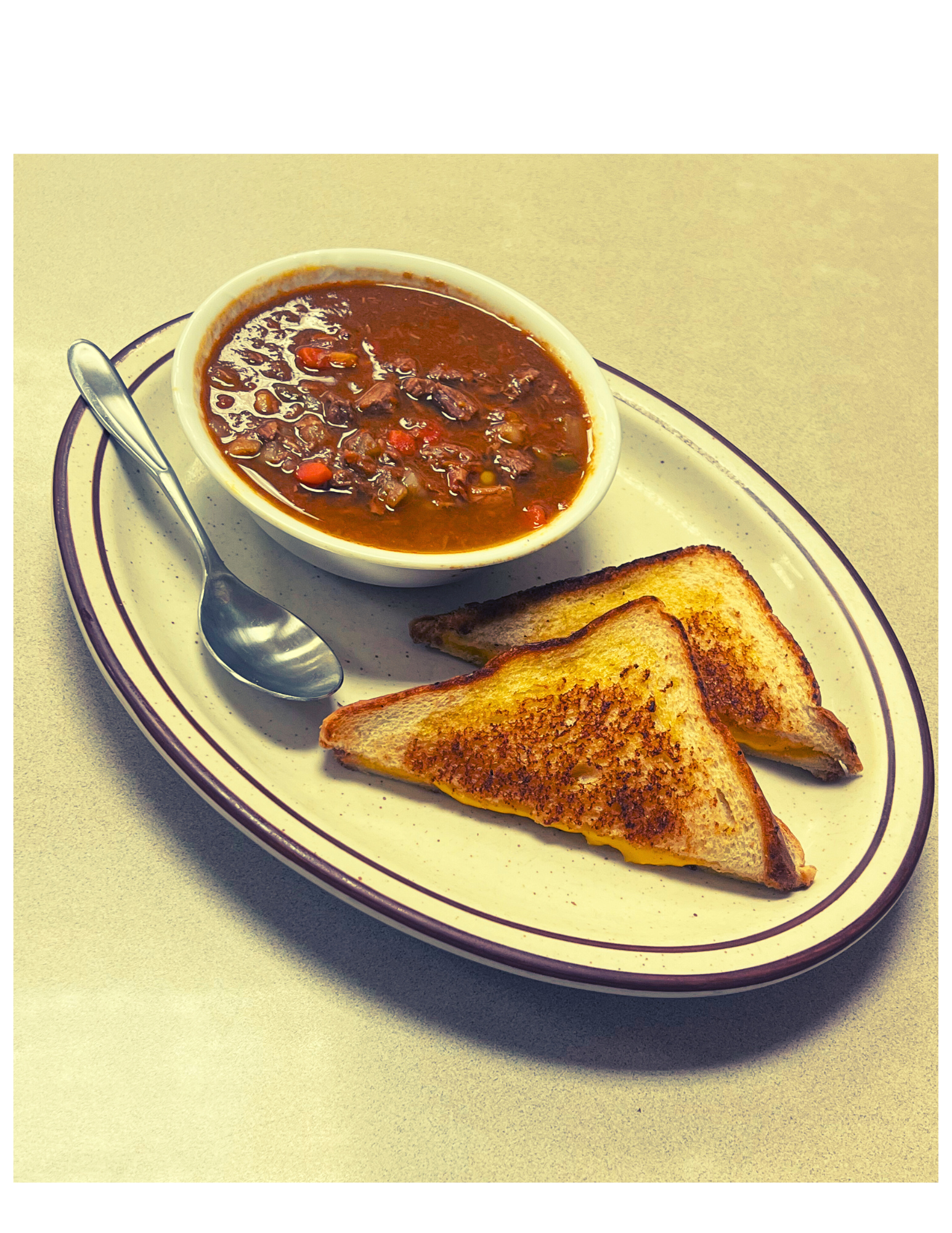 A plate with two toasted grilled cheese sandwiches, a bowl of vegetable beef soup, and a metal spoon on a light-colored table.