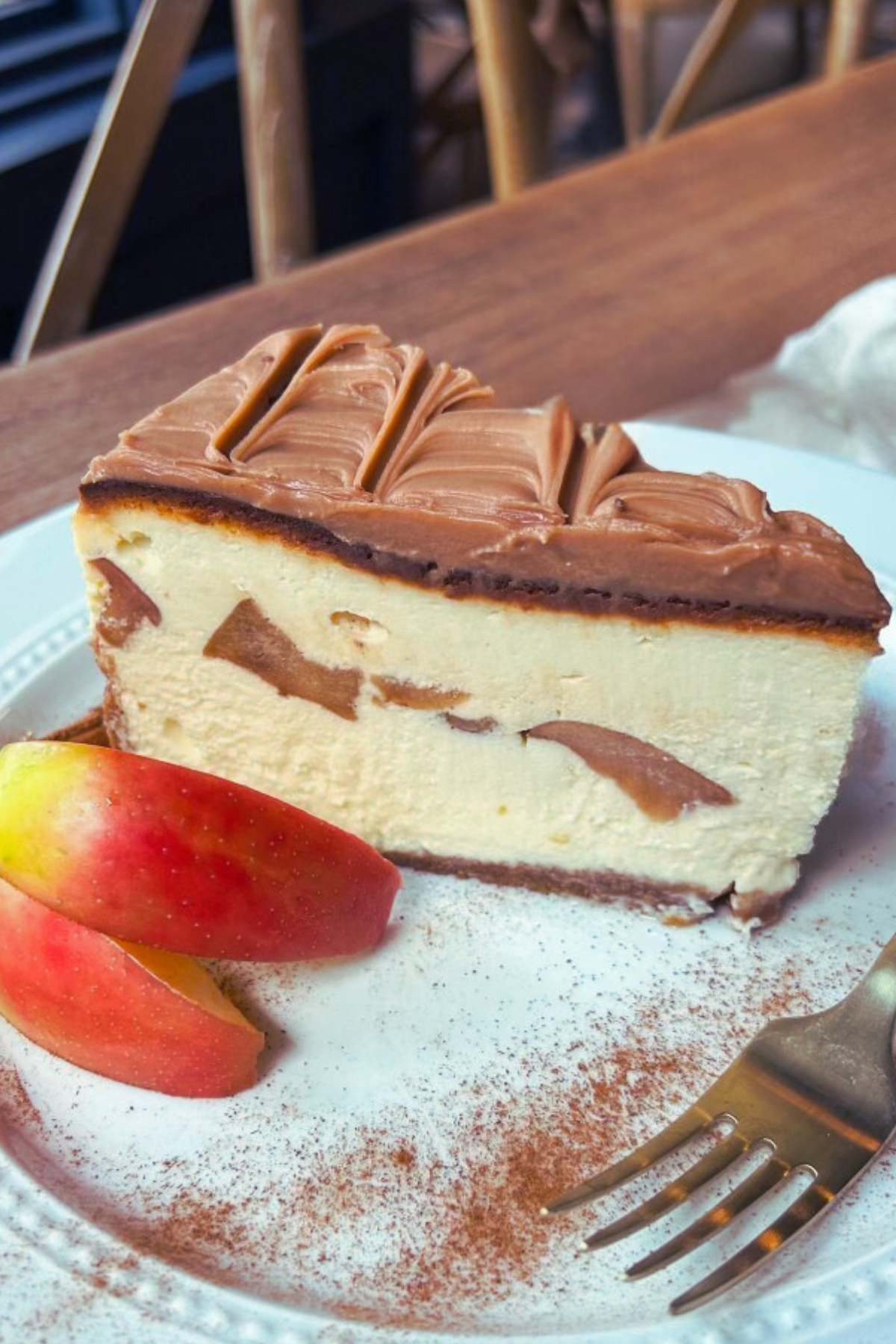 Slice of caramel apple cheesecake with caramel ganache and decorative cinnamon sugar, garnished with apple slices on a white plate