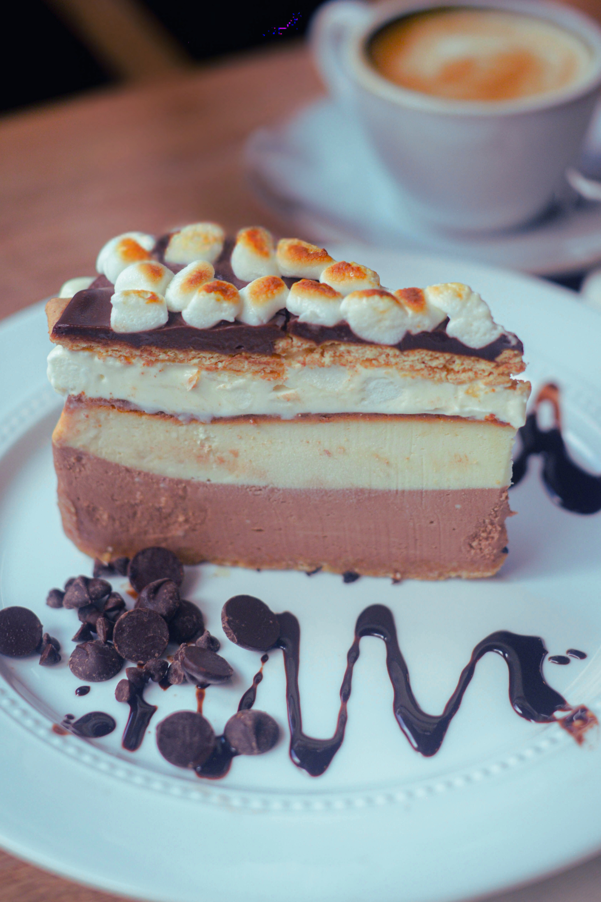 A slice of S'mores cheesecake with marshmallow frosting, graham crackers, chocolate ganache and marshmallows on top, served on a white plate with chocolate chips and chocolate sauce, with a cup of coffee in the background.