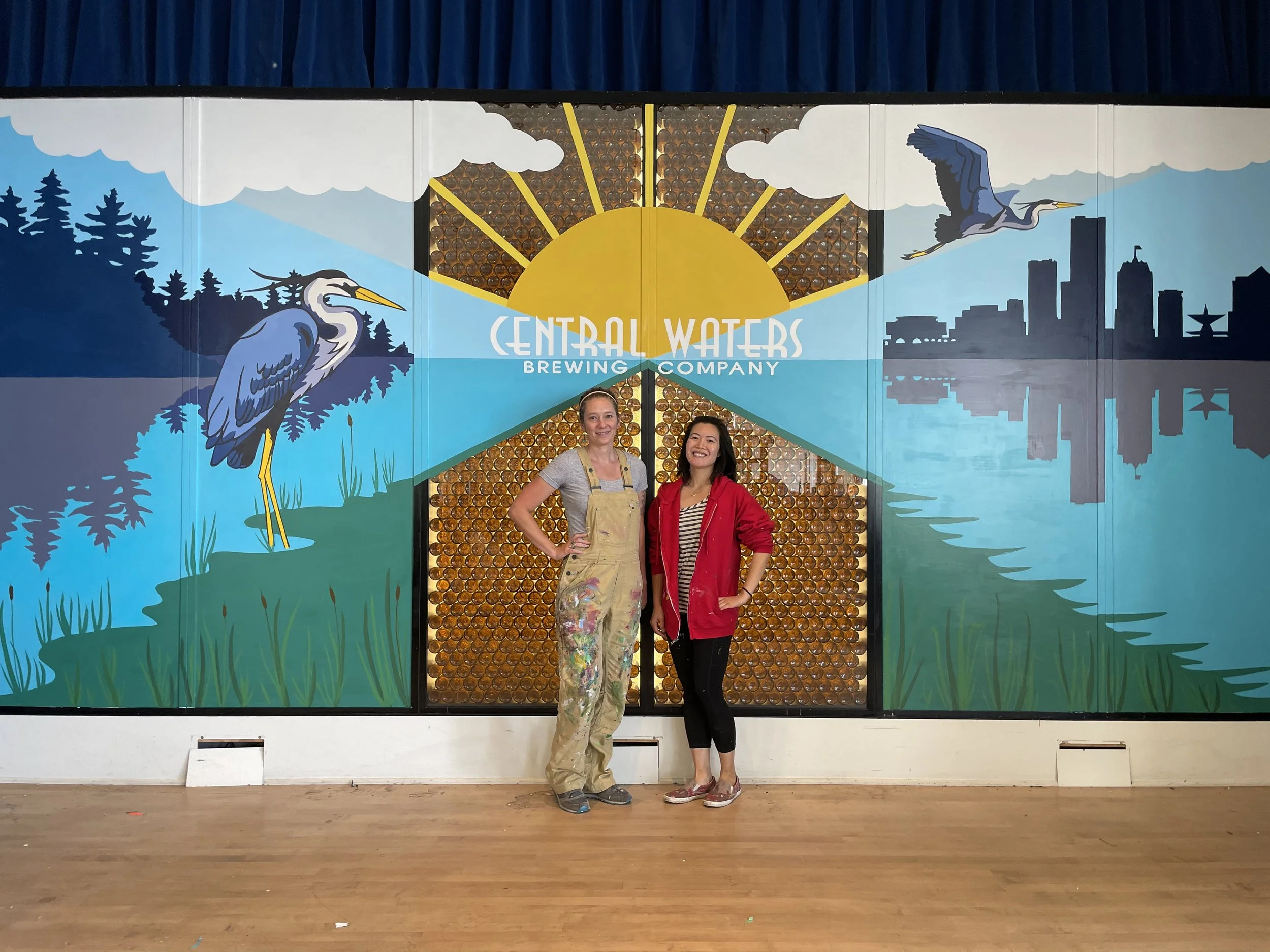 Two women standing in front of a mural for Central Waters Brewing Company, featuring a stylized sunset, herons, a city skyline, and water.