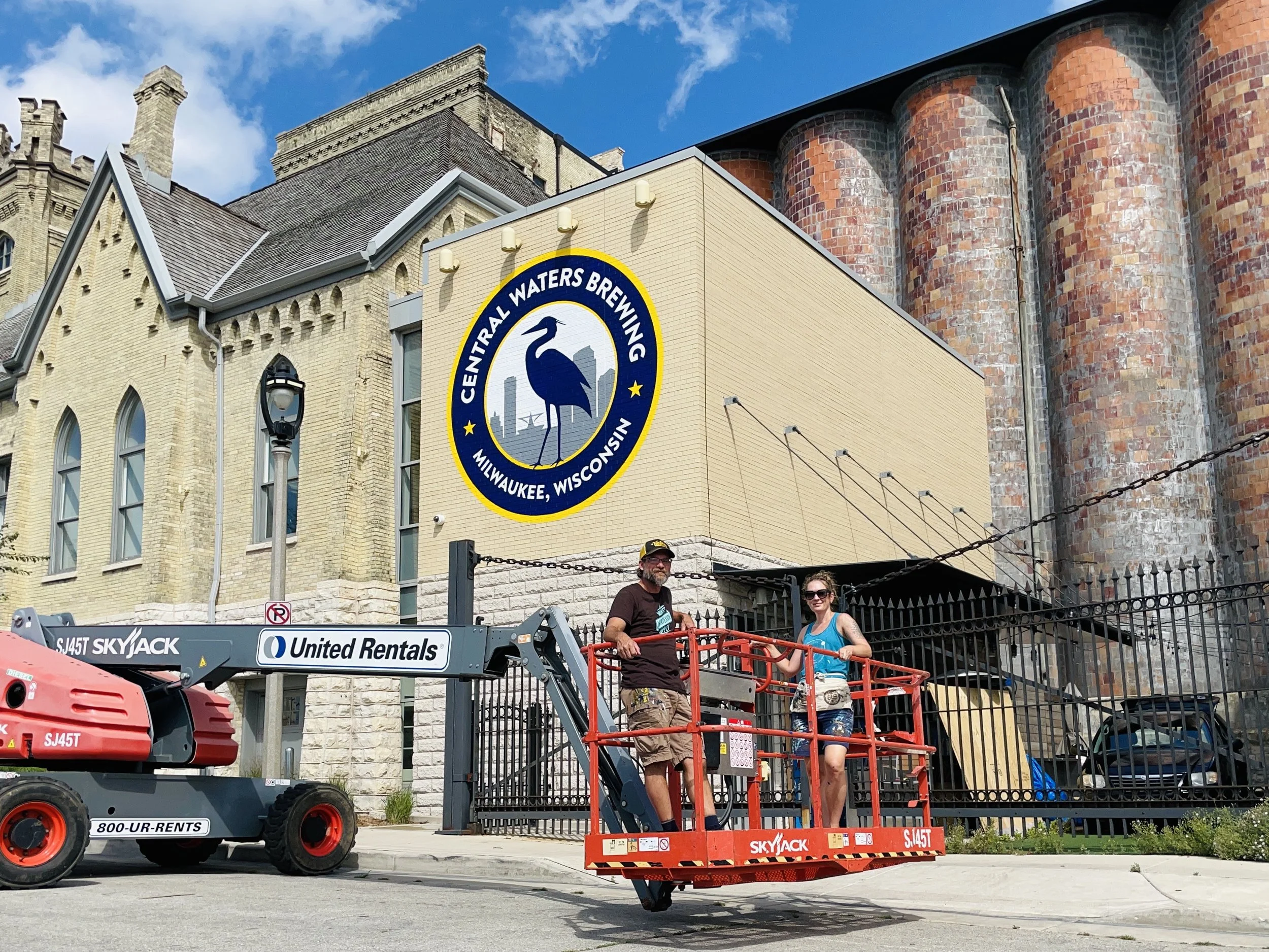 Two people on a lift platform near a building with a Wisconsin-themed sign reading 'Central Waters Brewing Milwaukee, Wisconsin.' Two large red silos are visible in the background.