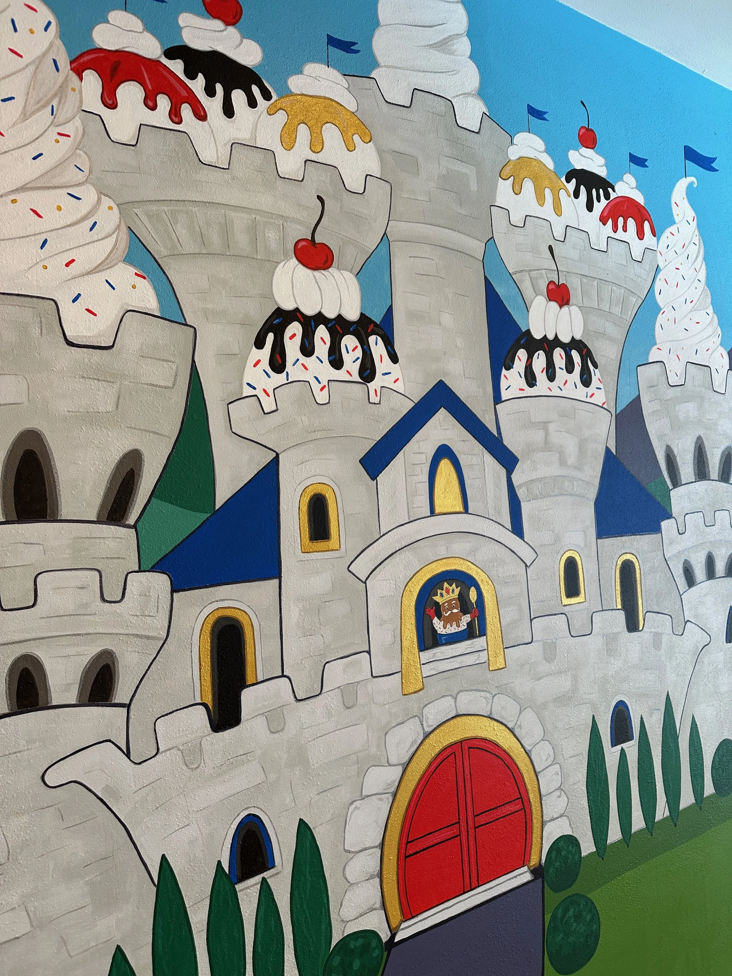 Colorful castle featuring towers with cupcake designs topped with cherries and drizzled with chocolate or icing, set against a blue sky.