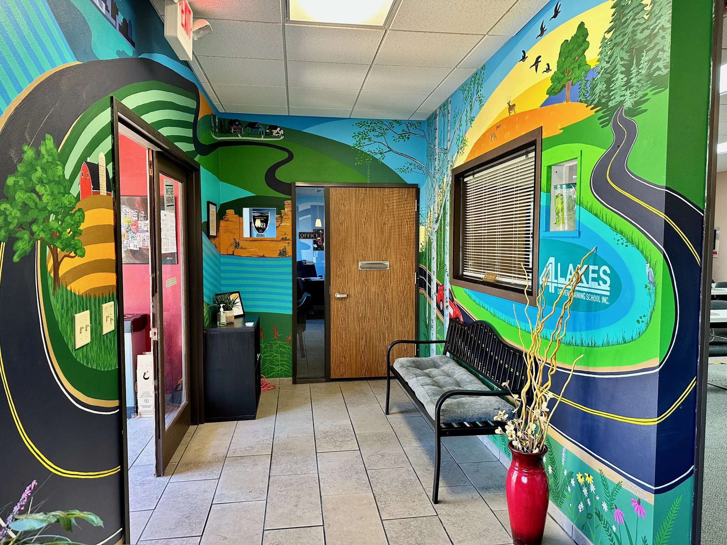 Colorful mural illustrating a winding road through natural landscapes with trees, hills, lakes, and wildlife in an office waiting area with a bench and decorative vase.