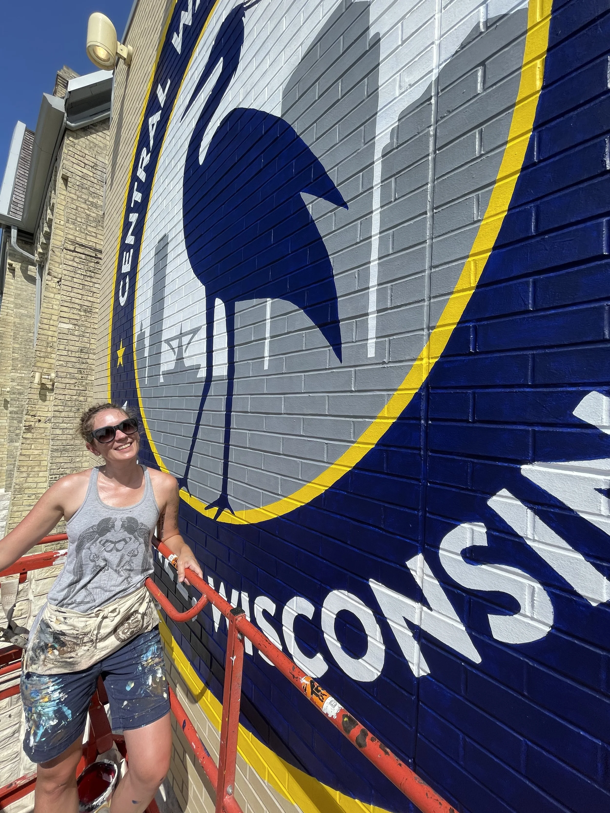 A woman smiling and wearing sunglasses, standing next to a cherry picker in front of a large mural of the Minnesota Vikings logo on a brick wall.