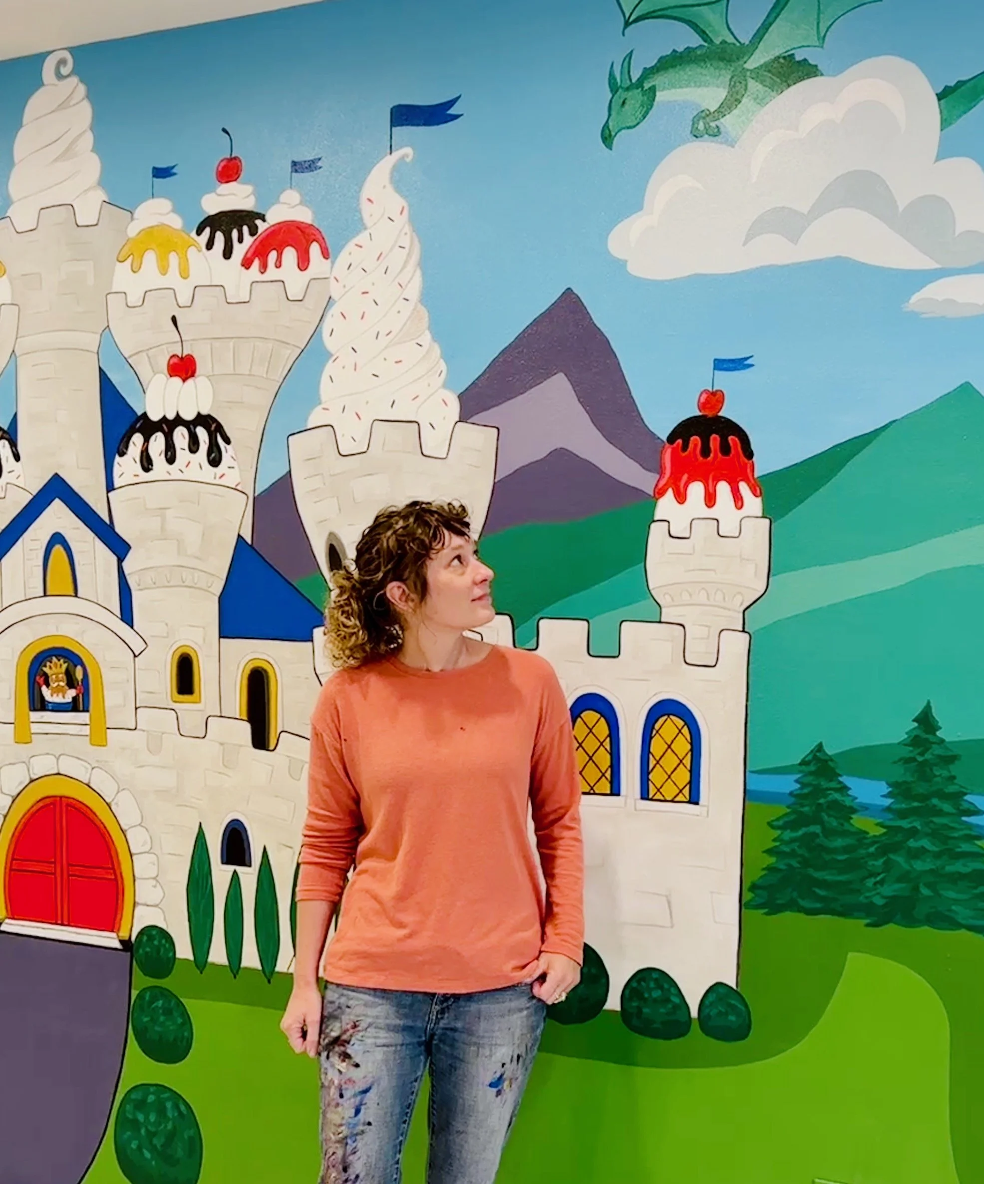 A woman standing inside a colorful mural of a fairytale castle with towers, mountains, clouds, trees, and dragons.