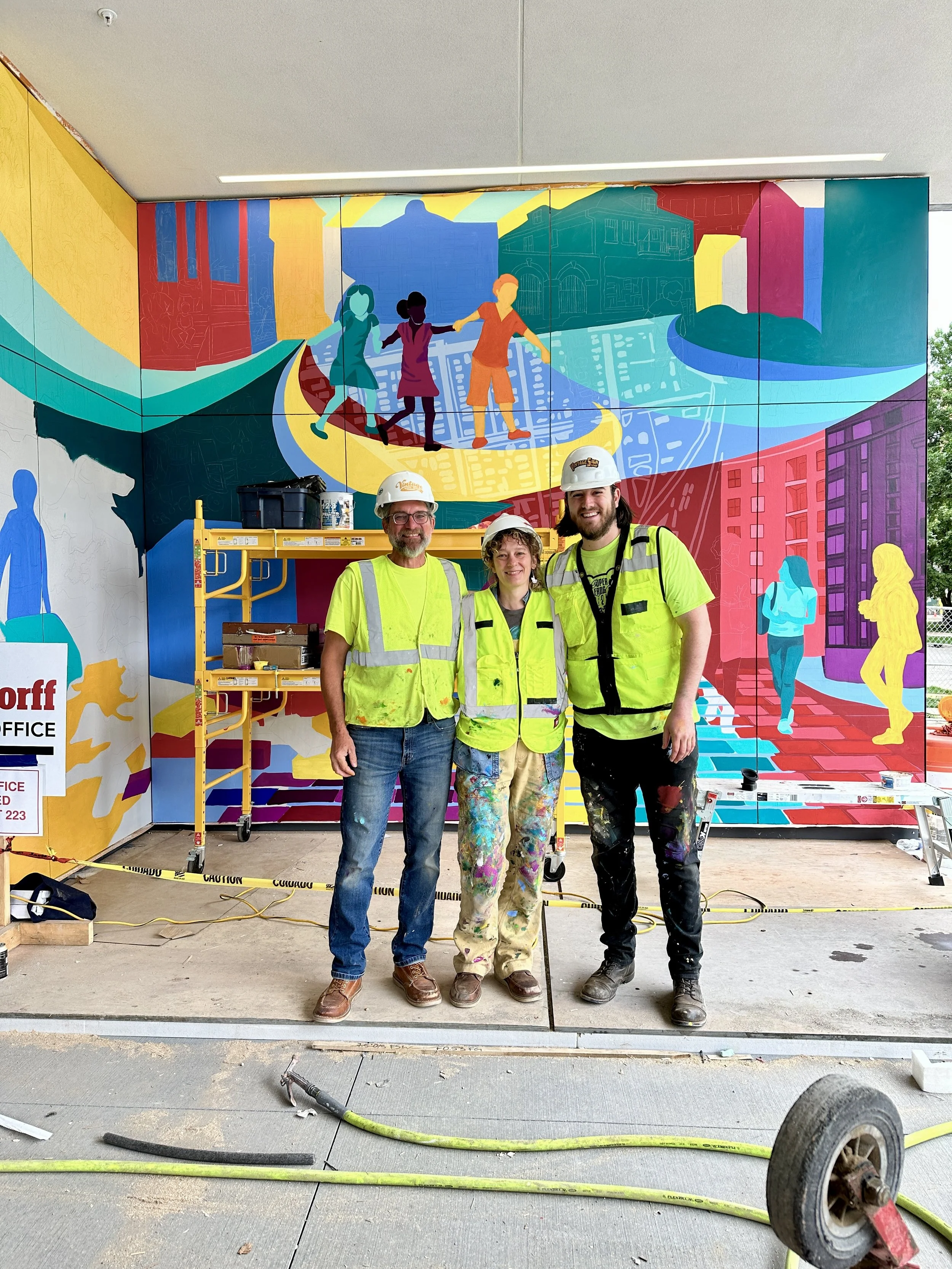 Three construction workers in yellow vests and hard hats standing in front of a colorful mural of children playing on a city street.