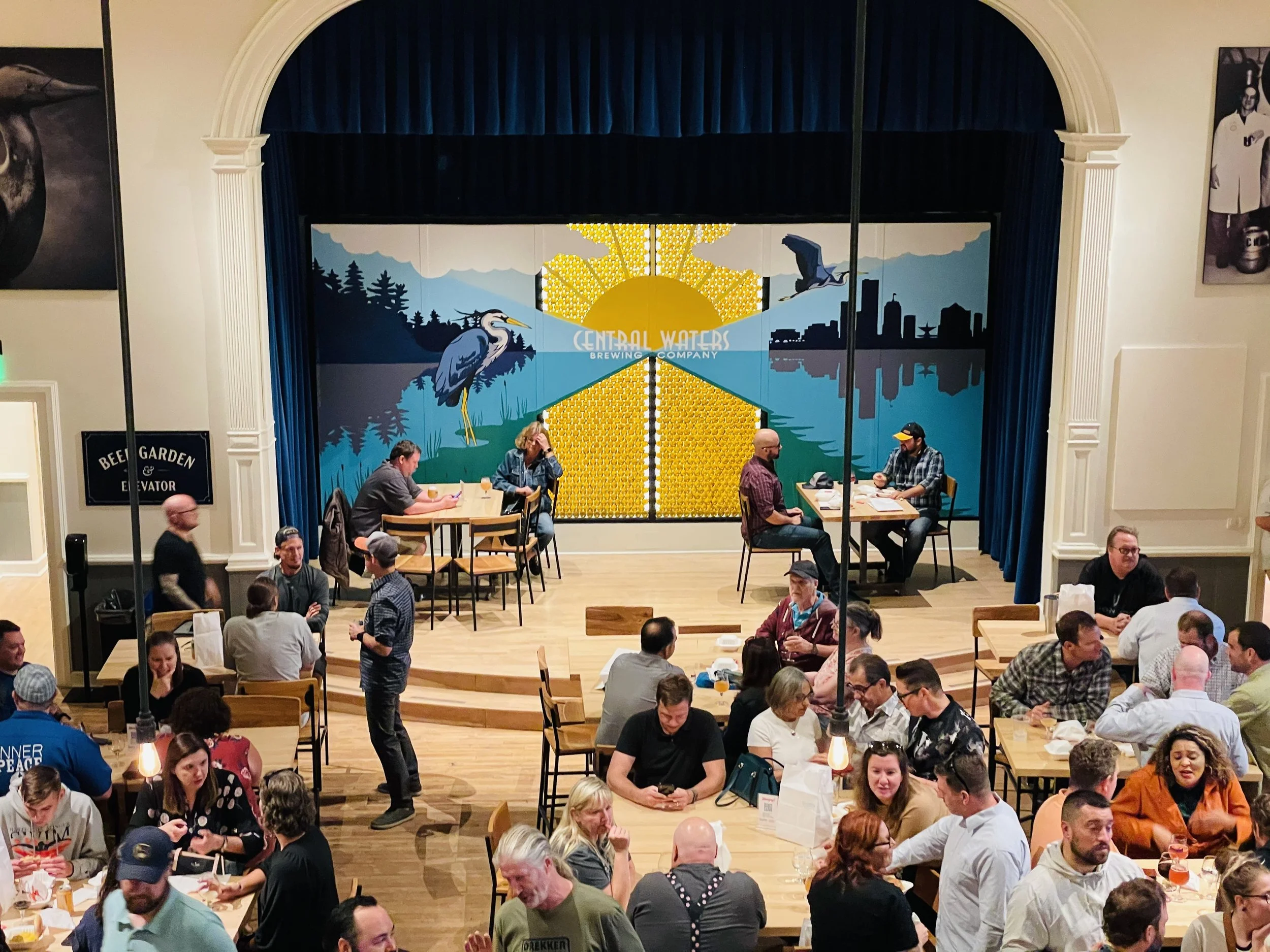 A crowded restaurant or beer hall with a stage at the front displaying a mural of a heron, trees, and city skyline, with the words "Central Waters Brewing Company". People are sitting at tables, dining, talking, and socializing.
