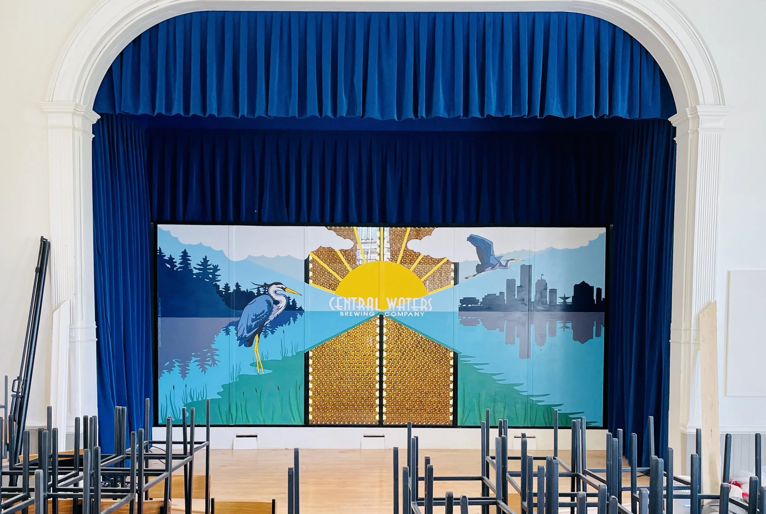 Stage with a colorful mural of a heron, city skyline, and sunrise, with blue curtains and stacked chairs in front.