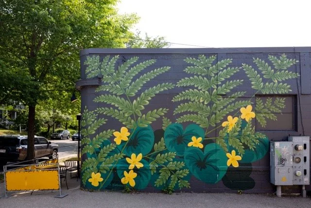 Colorful mural of green ferns and yellow flowers on the side of a building, with trees and parked cars in the background.