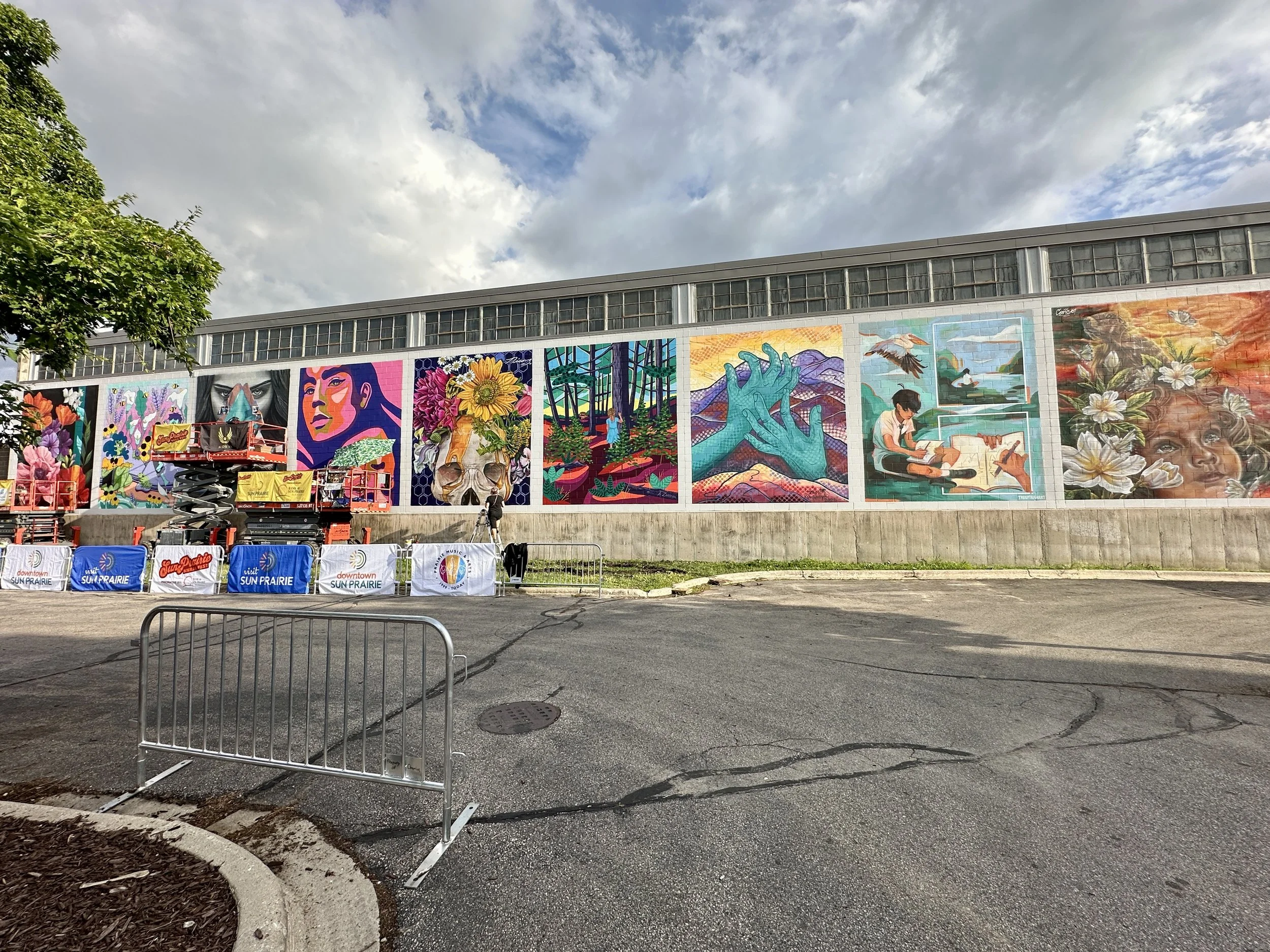 A large wall mural with various colorful artwork, including portraits, nature scenes, and abstract designs, being prepared for display with scaffolding and barriers in the foreground.