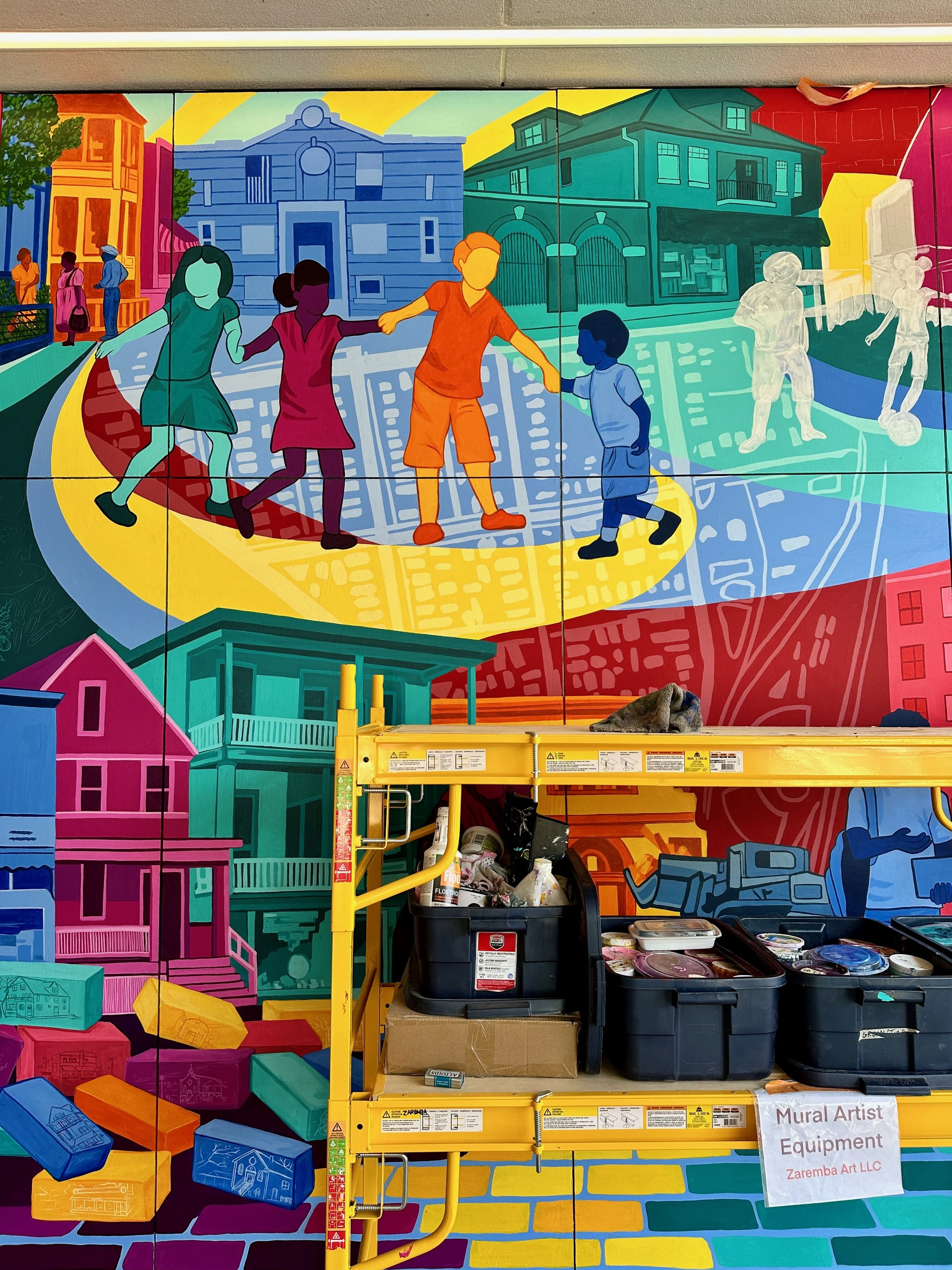 Colorful mural of children playing hopscotch and soccer on a city street, with vibrant houses and buildings in the background, under a yellow sky. Art supplies and equipment are on a yellow scaffold in the foreground.
