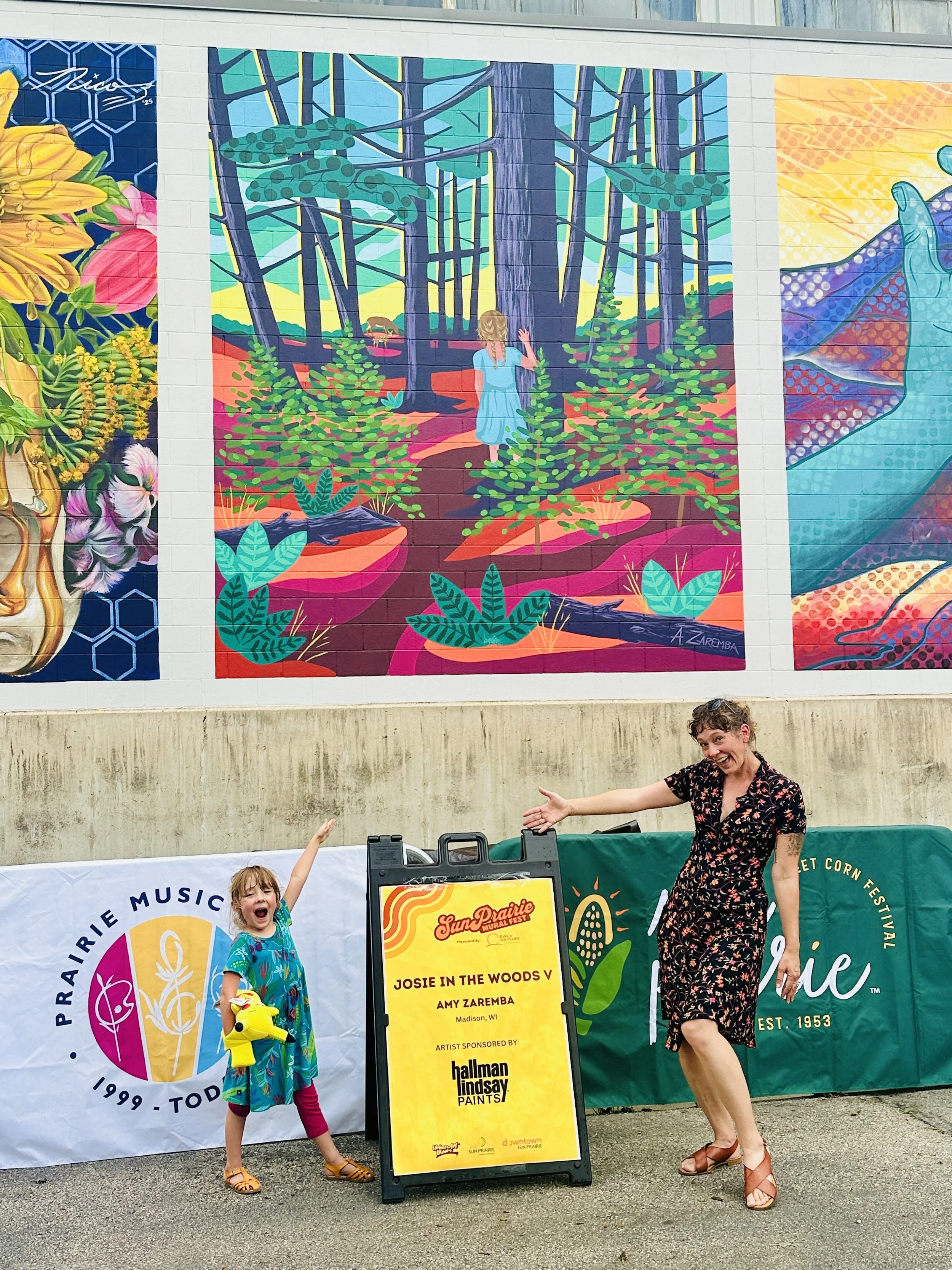 A woman and a young girl posing in front of colorful wall murals at the Prairie Music Festival. The girl holds a yellow toy, and both are smiling. A sign in front displays the concert details.