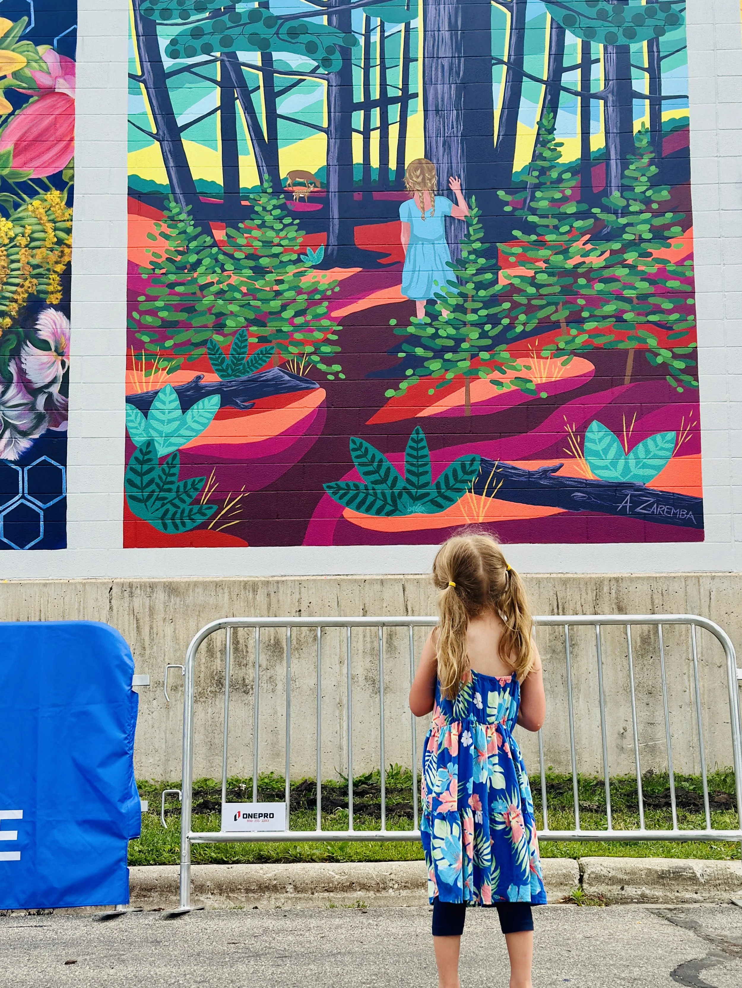 A young girl with long hair in pigtails, wearing a colorful floral dress and dark leggings, stands in front of a vibrant wall mural of a forest scene with trees, bushes, and a girl in a blue dress waving. She is behind metal barricades on a sidewalk.