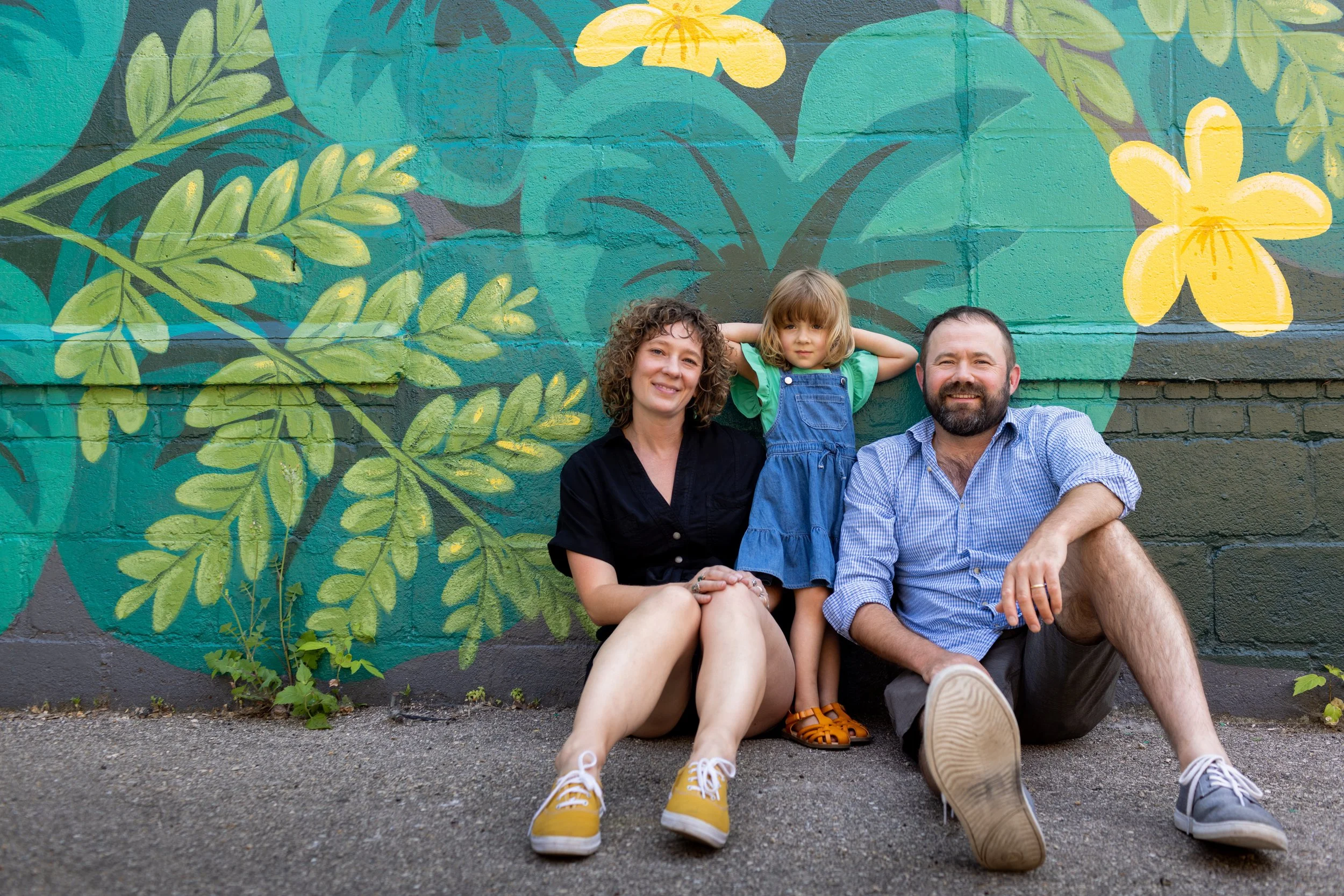 A family of three sitting on the ground in front of a colorful mural with tropical leaves and yellow flowers, including a woman with curly hair in a black shirt, a young girl with blond hair in a blue dress and green shirt, and a man with a beard in 
