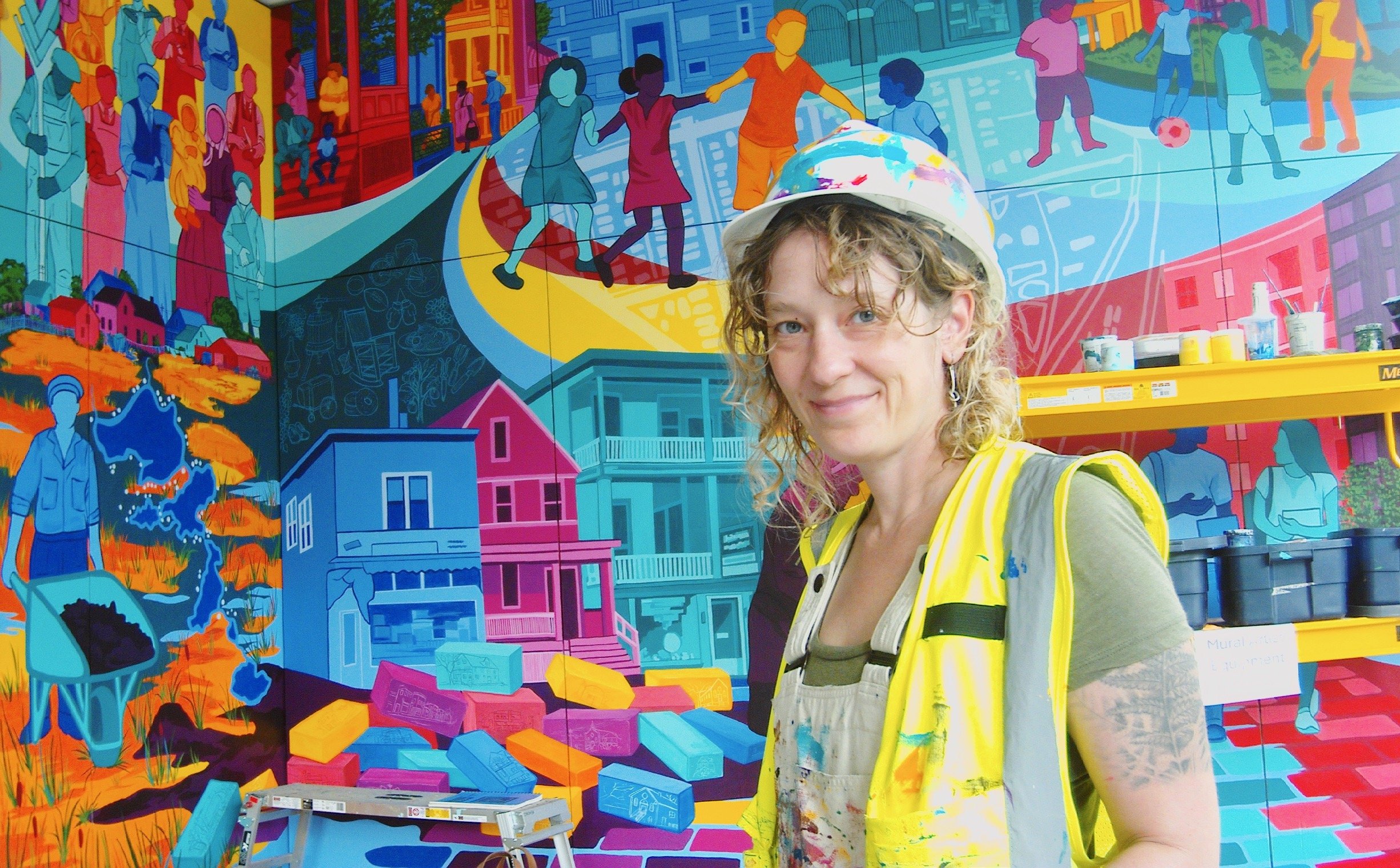 Woman in a yellow safety vest and a white painter's hat stands in front of a colorful mural depicting various scenes of community life, including children playing, houses, and people working.