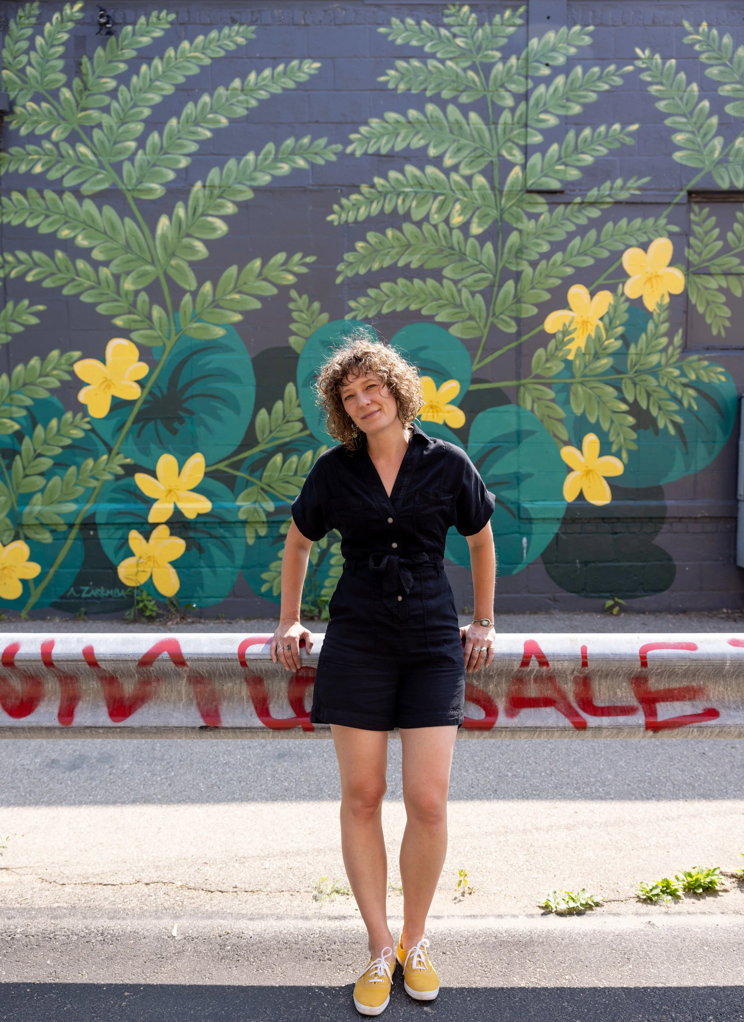 A woman with curly hair wearing a black dress and yellow sneakers standing in front of a colorful mural of tropical leaves and yellow flowers on a gray brick wall.