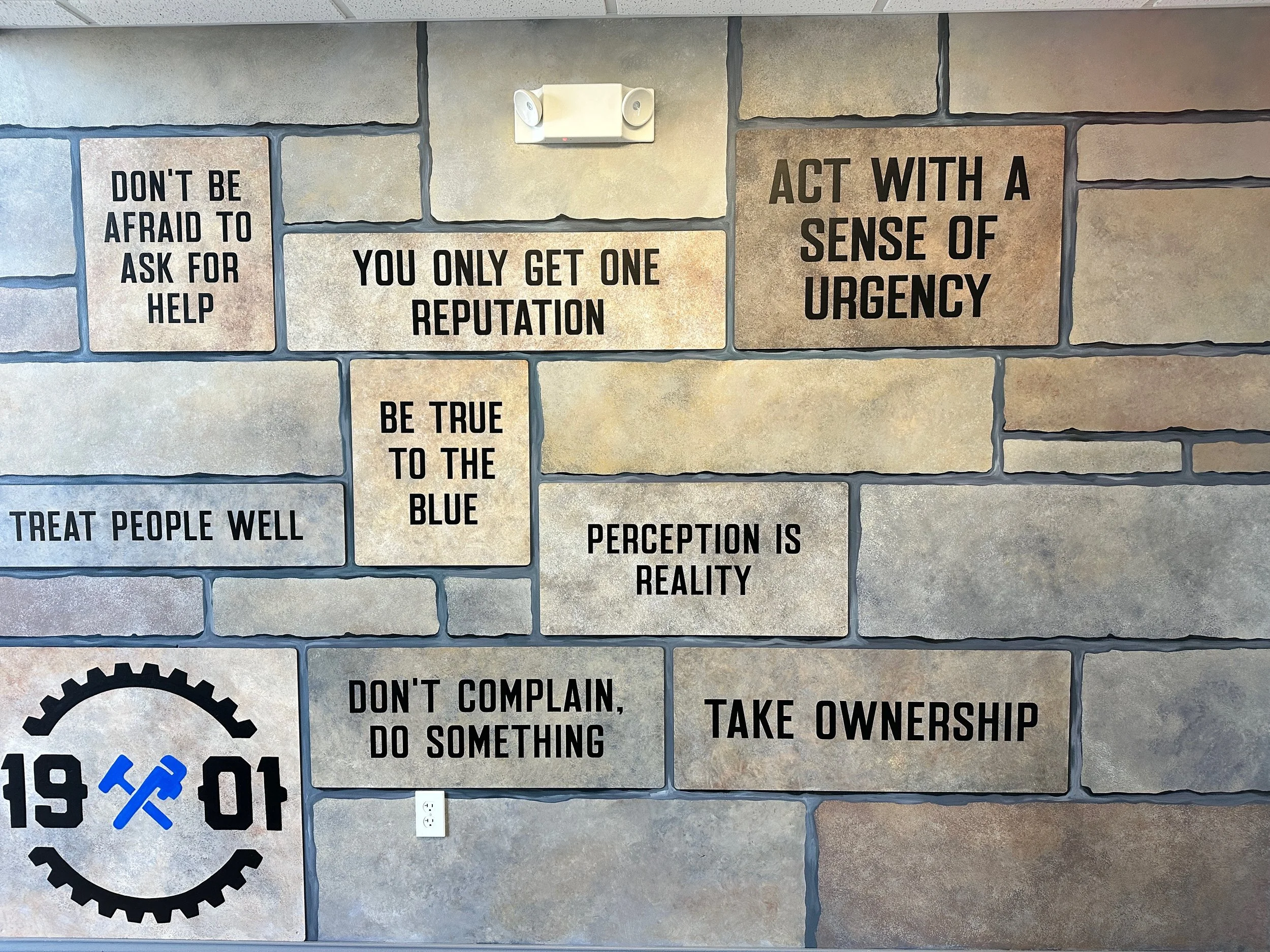 Wall with motivational quotes on stone tiles, including phrases like 'Don't be afraid to ask for help,' 'You only get one reputation,' 'Act with a sense of urgency,' 'Be true to the blue,' 'Perception is reality,' 'Treat people well,' 'Don't complain