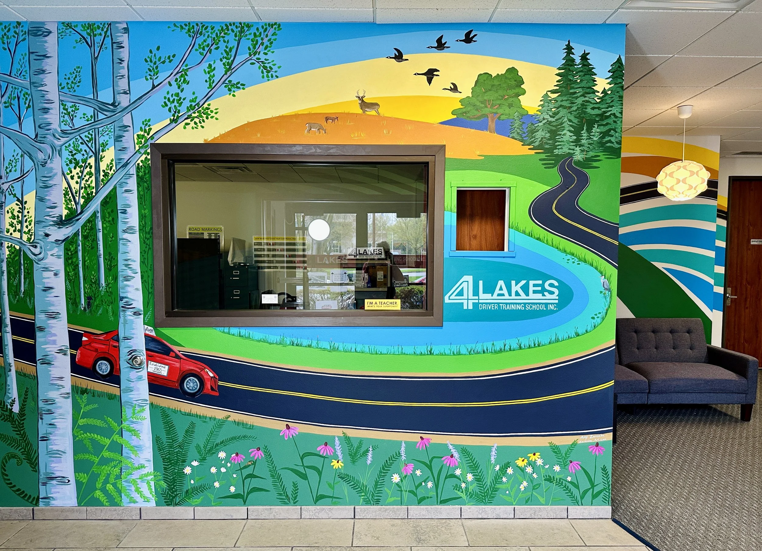 Colorful wall mural at a driver training school depicting a scenic landscape with trees, a river, a sunset, wildlife, a road, and a red car, with windows and a door in the wall, and a gray sofa nearby.