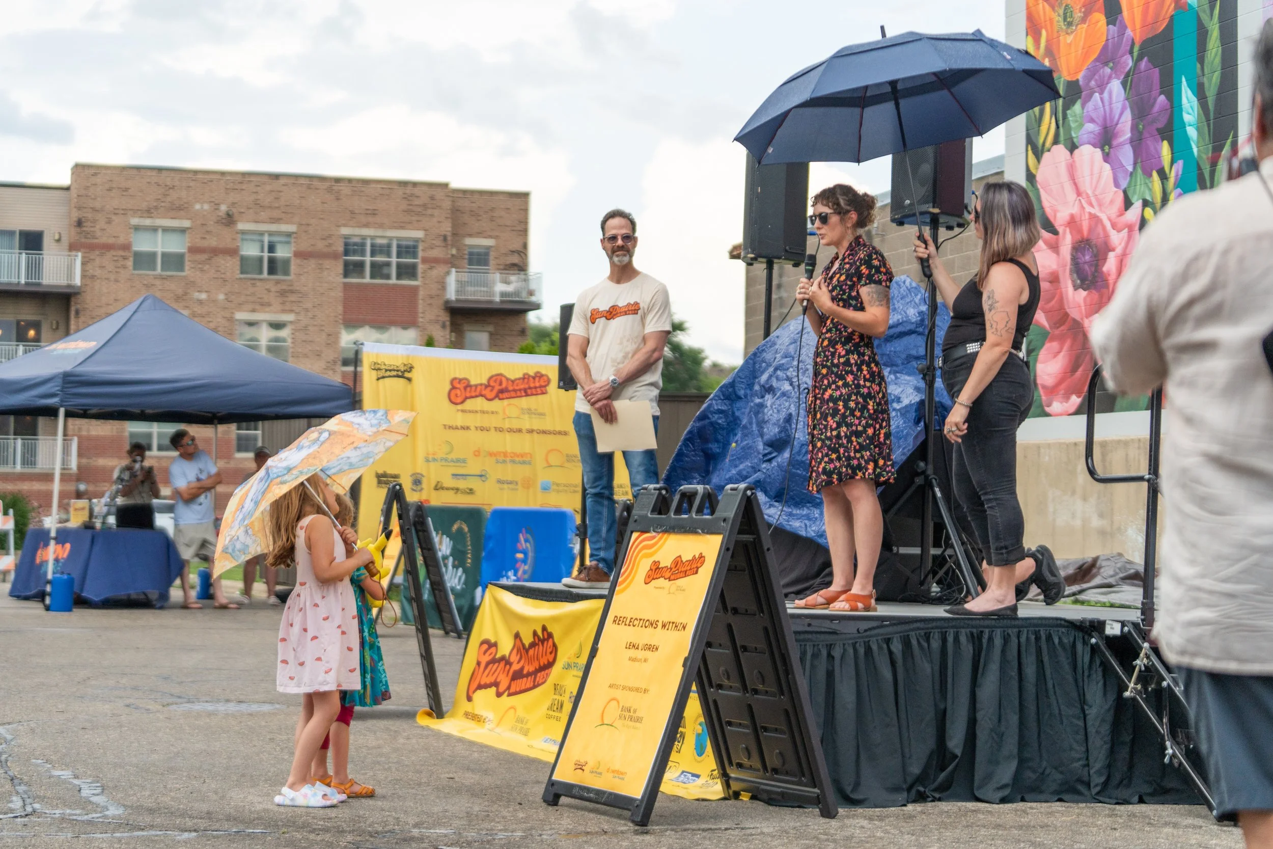 People attending an outdoor event, with a woman speaking into a microphone on stage, a man standing next to her, and a girl with an umbrella in the foreground.