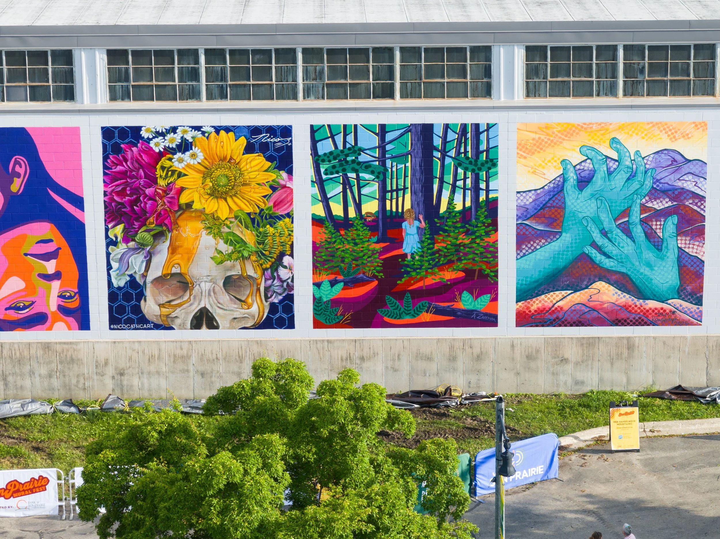 Colorful mural paintings on a wall, including abstract faces, a skull decorated with flowers, a girl in a forest, and blue hands over mountains.
